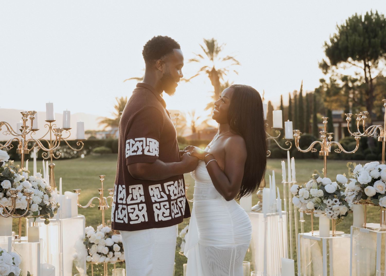 A couple sharing a romantic moment together at a candlelit outdoor proposal setup at sunset.
