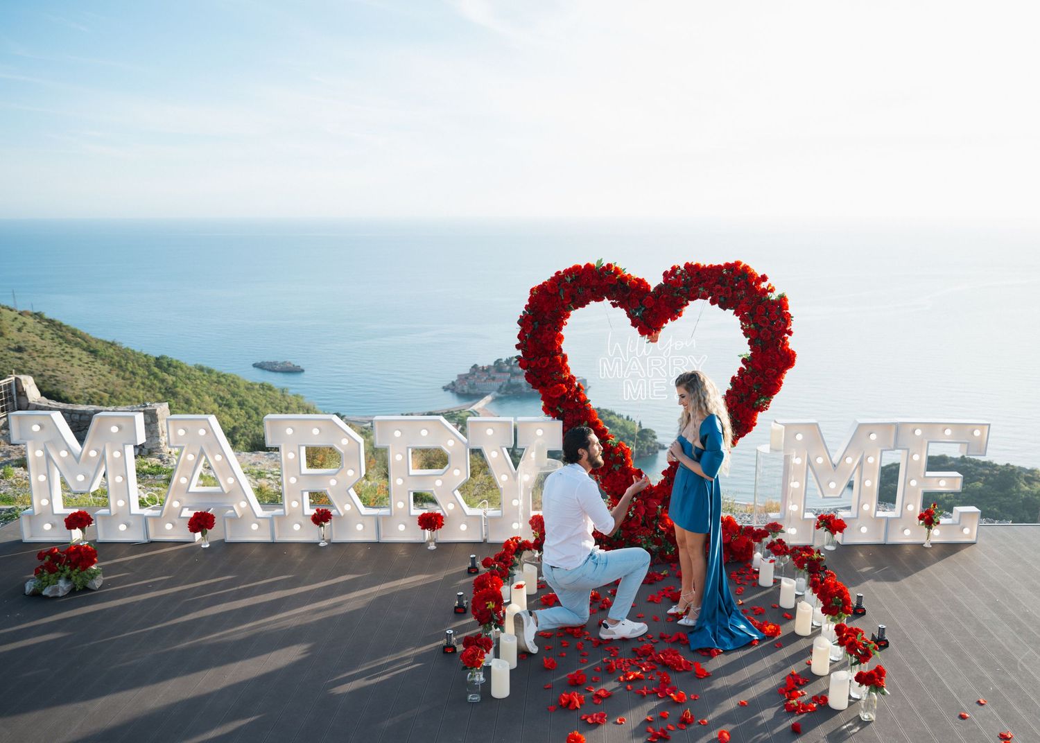 Man on one knee proposing to woman under red rose heart arch with large 'Marry Me' letters by the sea.