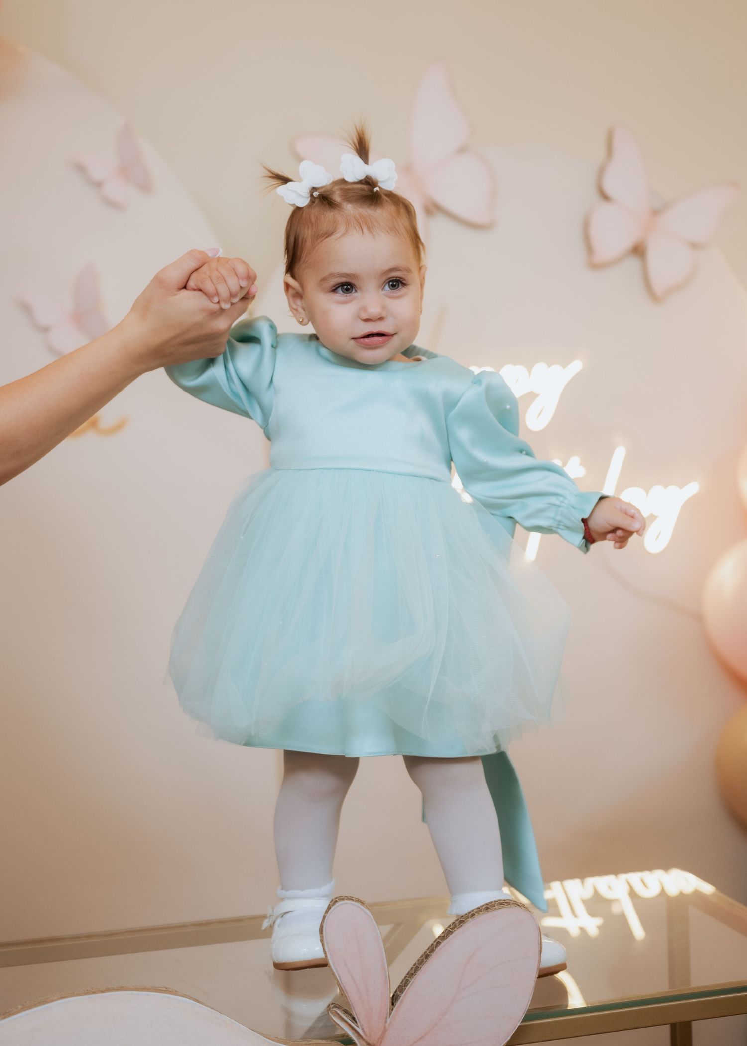 A toddler in a mint tulle dress stands with assistance at a butterfly-themed birthday party celebration.
