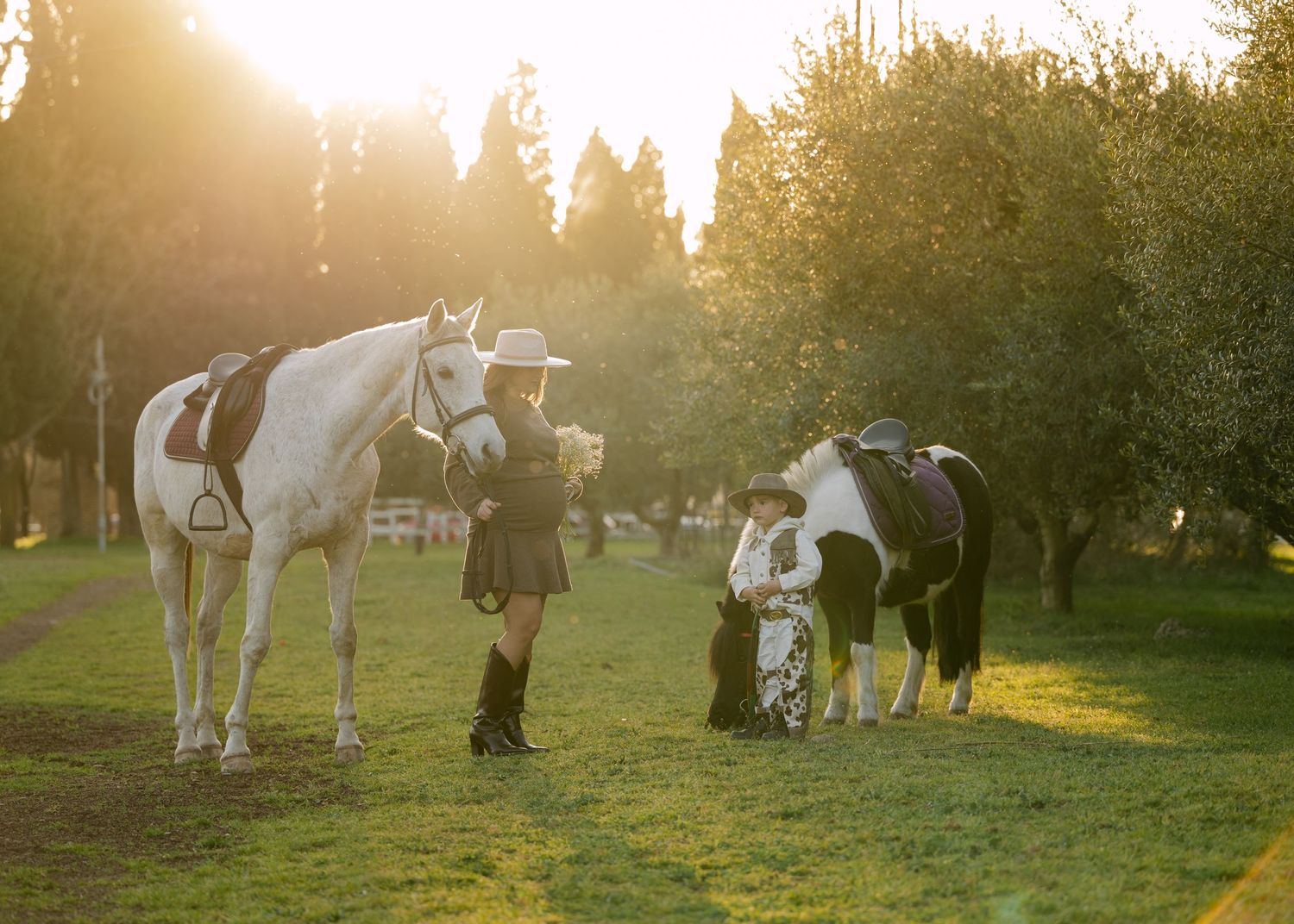 A pregnant woman in a hat stands with two horses on a sunlit green field surrounded by trees.