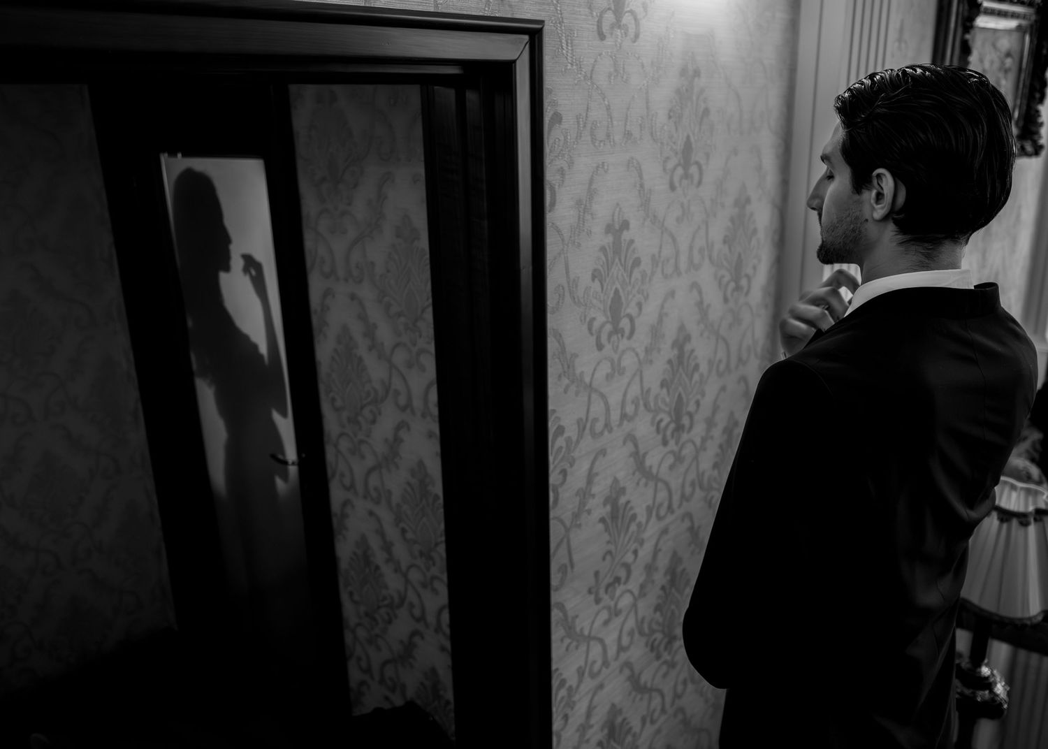 Black and white photo of groom in suit adjusting tie while bride's silhouette reflects in mirror.