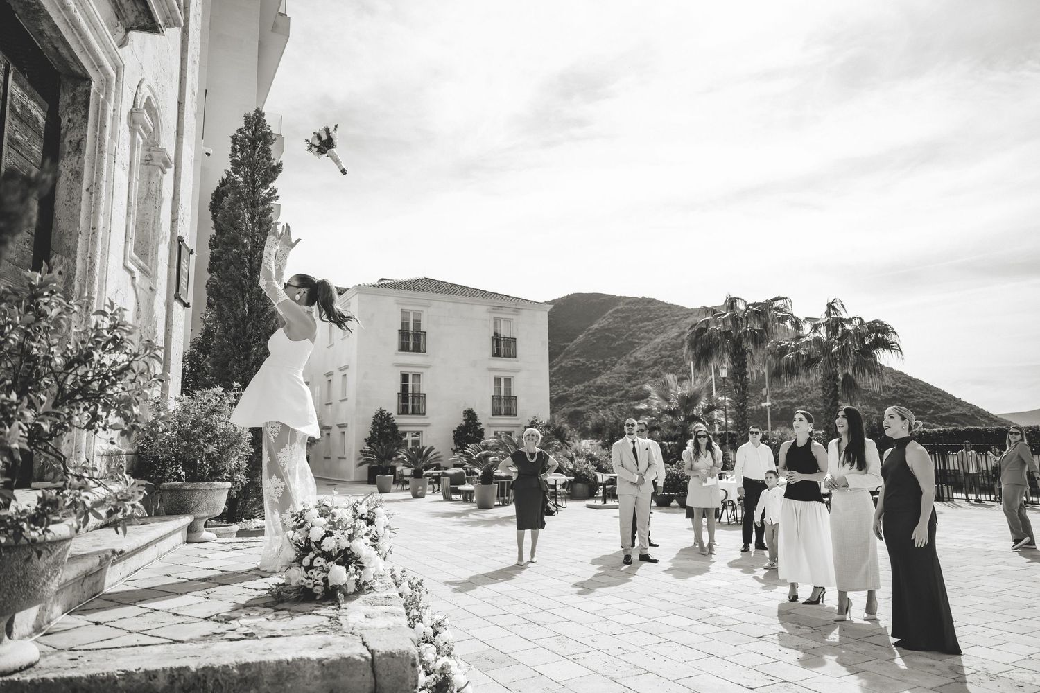Bride tosses bouquet to guests on elegant outdoor terrace with mountain backdrop at wedding celebration.