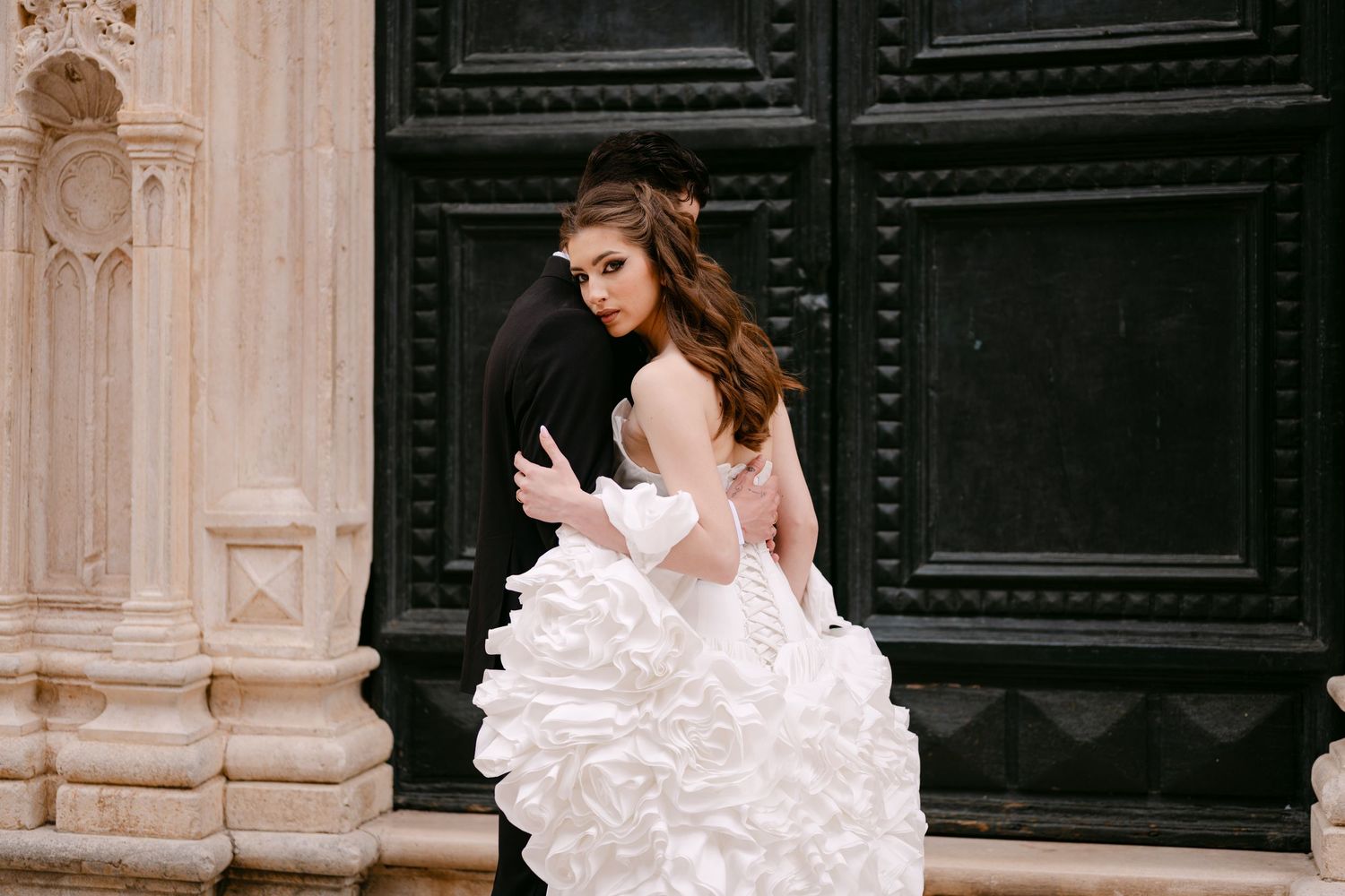 Bride in dramatic ruffled ball gown embraces groom against grand dark wooden doors of historic building.