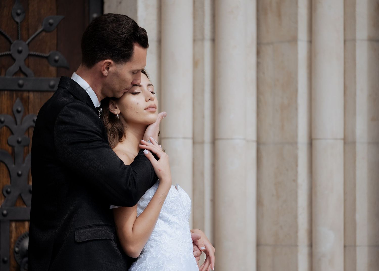 Groom tenderly embracing bride from behind against tall stone columns at a historic venue.