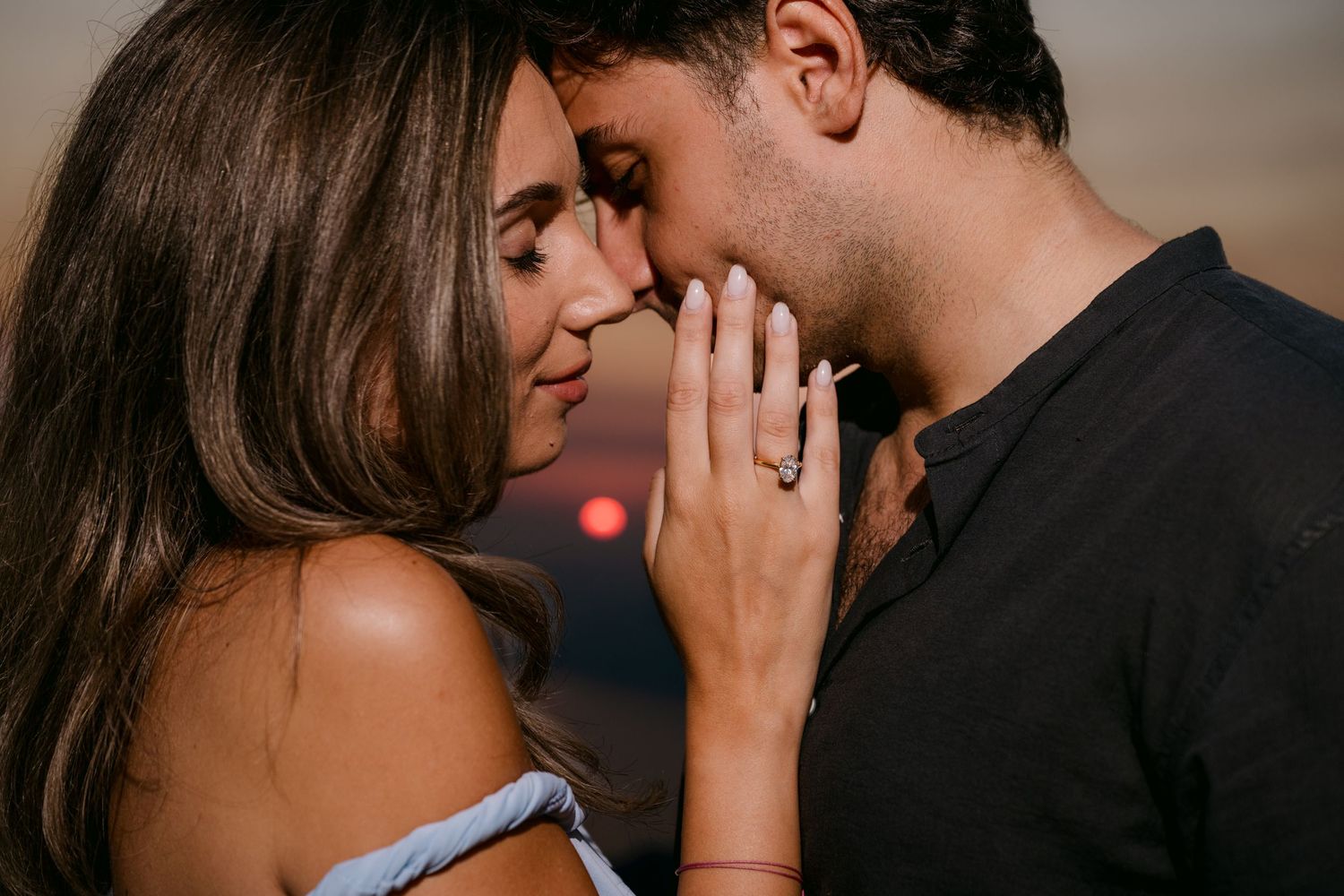 Intimate close-up of an engaged couple touching foreheads at sunset, woman showing diamond ring.