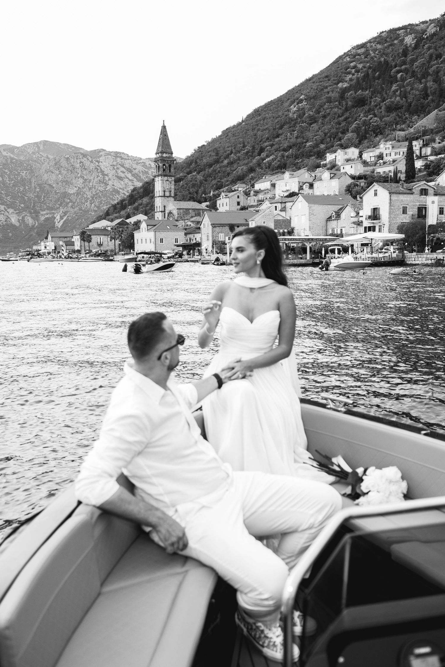 Black and white photo of newlyweds on a boat with Perast church tower and Bay of Kotor coastline behind.