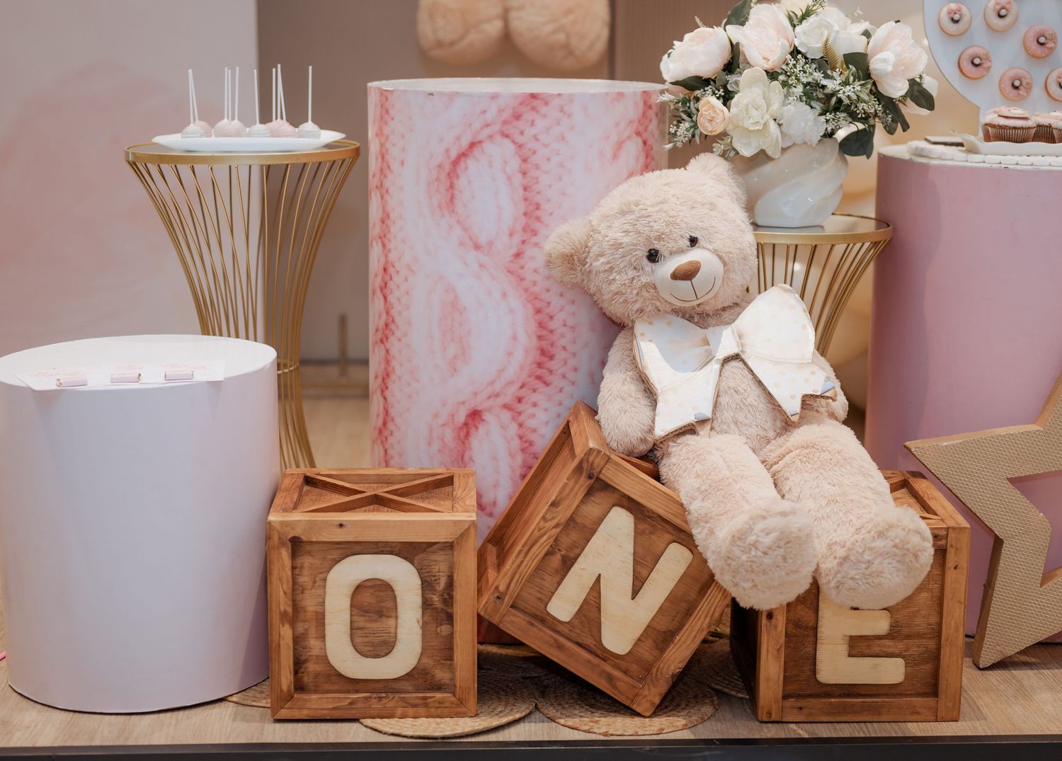 Plush teddy bear posed on wooden ONE letter blocks with pink birthday party decor nearby.