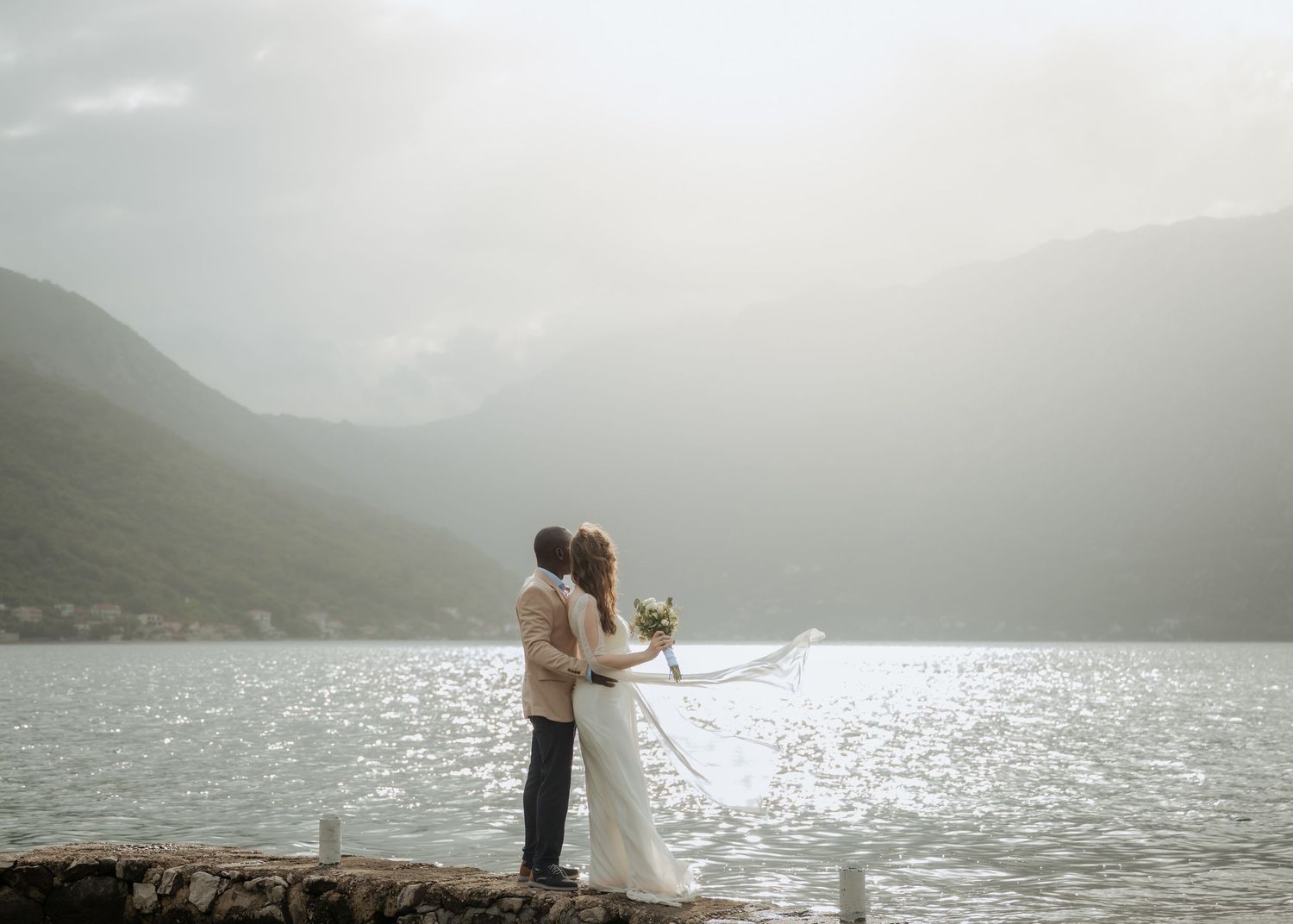 Bride and groom share a kiss by a serene mountain lake with veil flowing in the breeze.