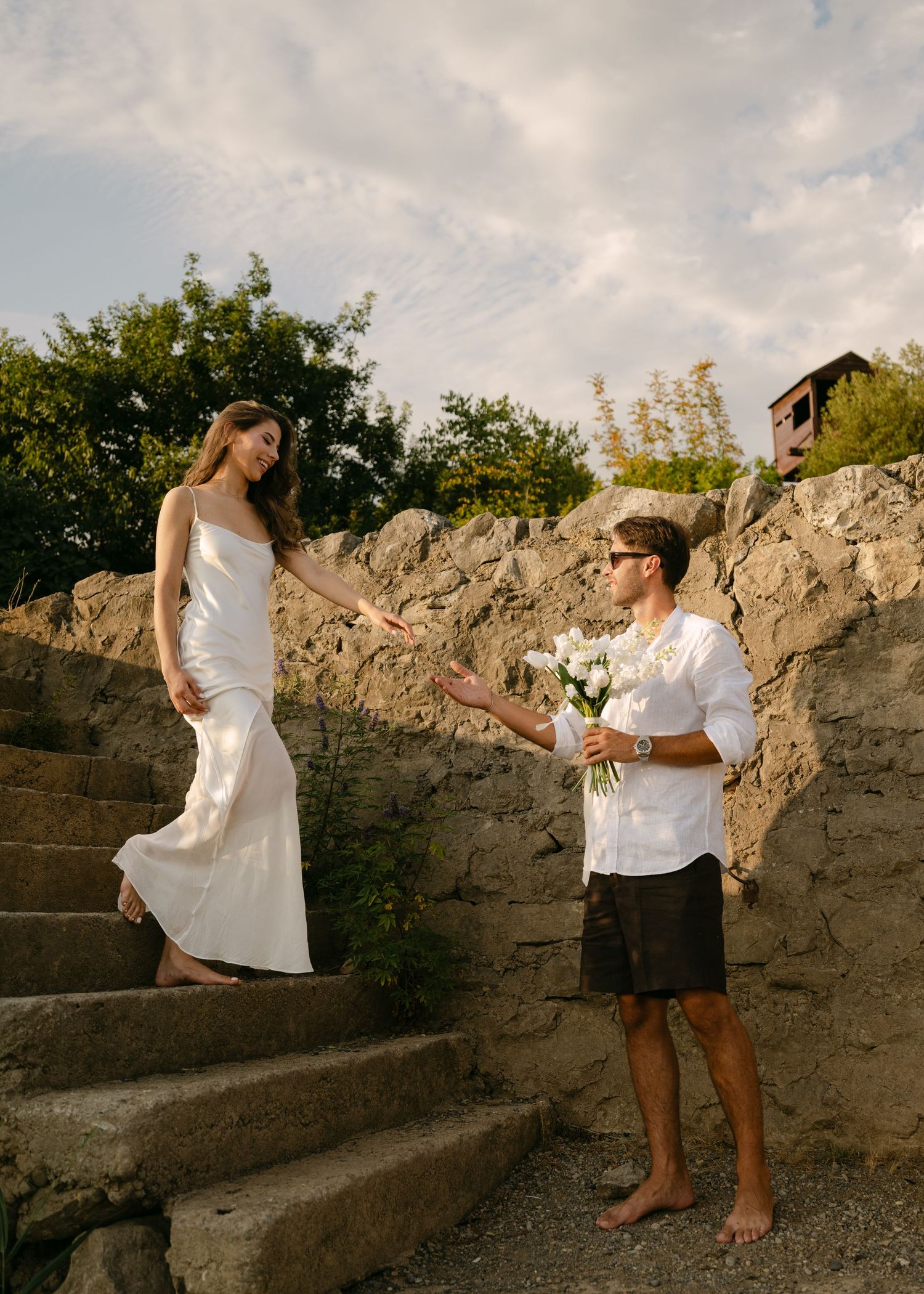 Groom offering white flower bouquet to bride in white dress on rustic stone steps outdoors.
