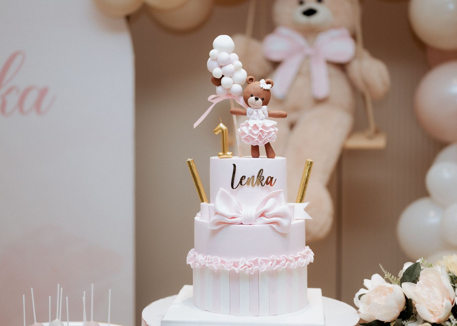 Two-tier pink birthday cake named Lenka with a dancing bear topper and gold number one candle.