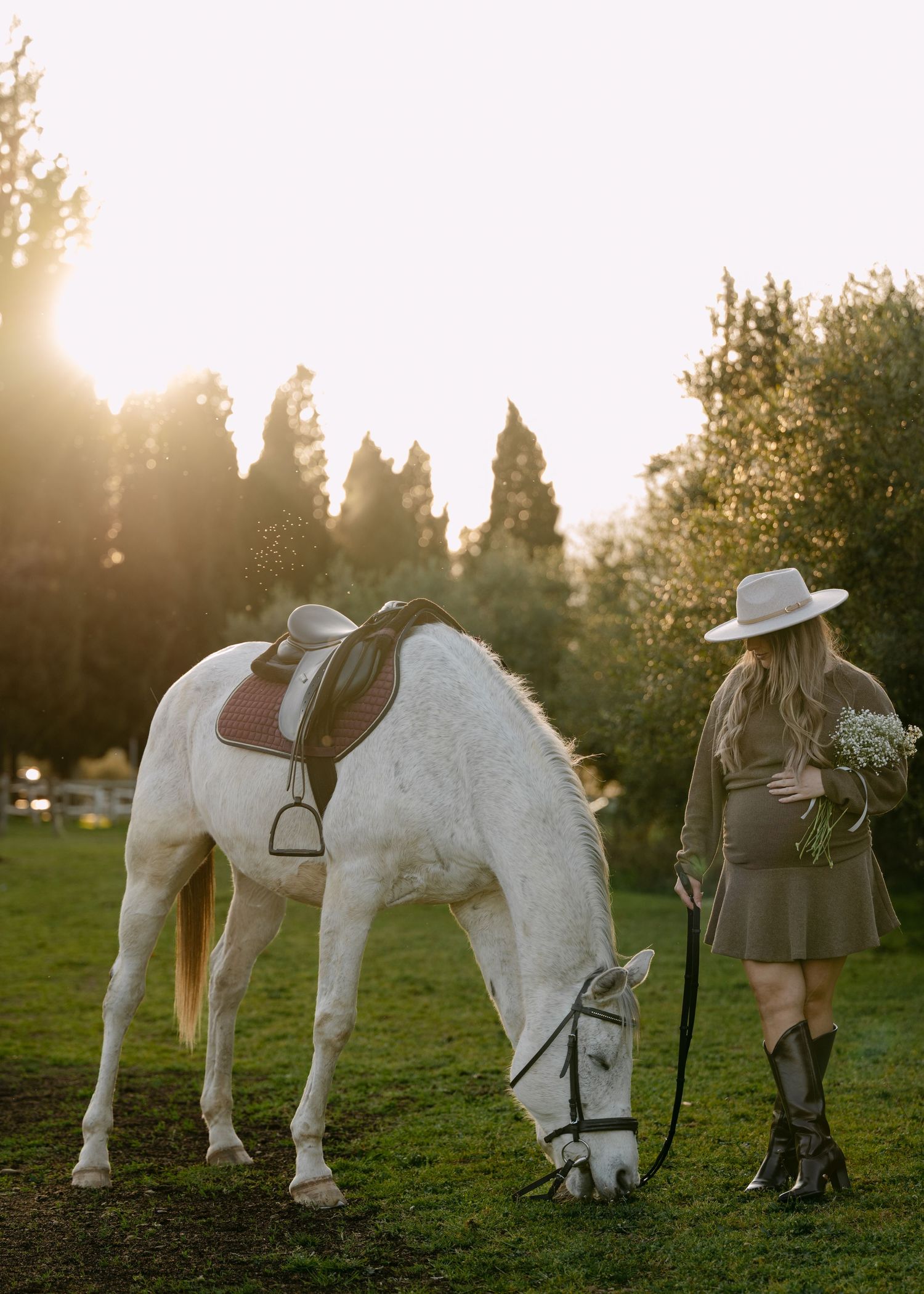 A pregnant woman in a hat and boots stands beside a white grazing horse during a golden sunset.