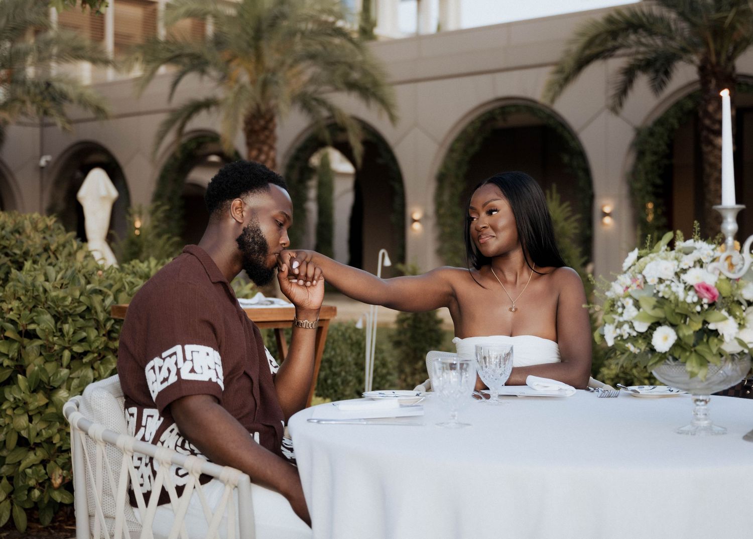 A couple enjoys a romantic outdoor dinner, the woman extending her hand across a white tablecloth.