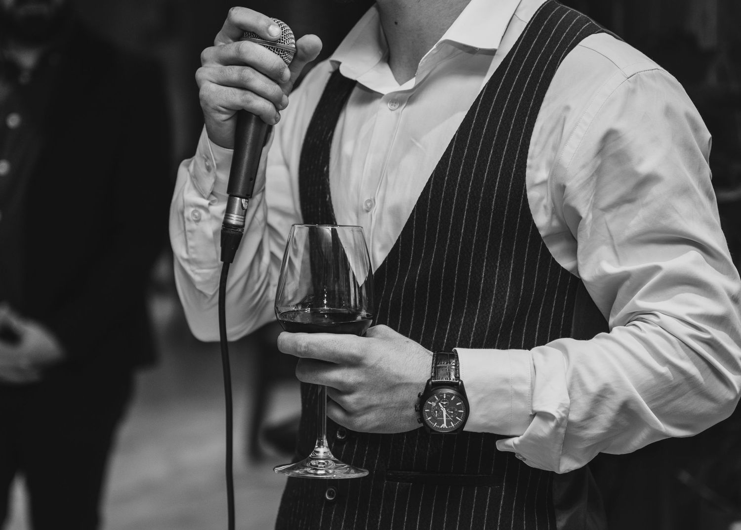 Man in pinstripe vest holding microphone and glass of red wine.