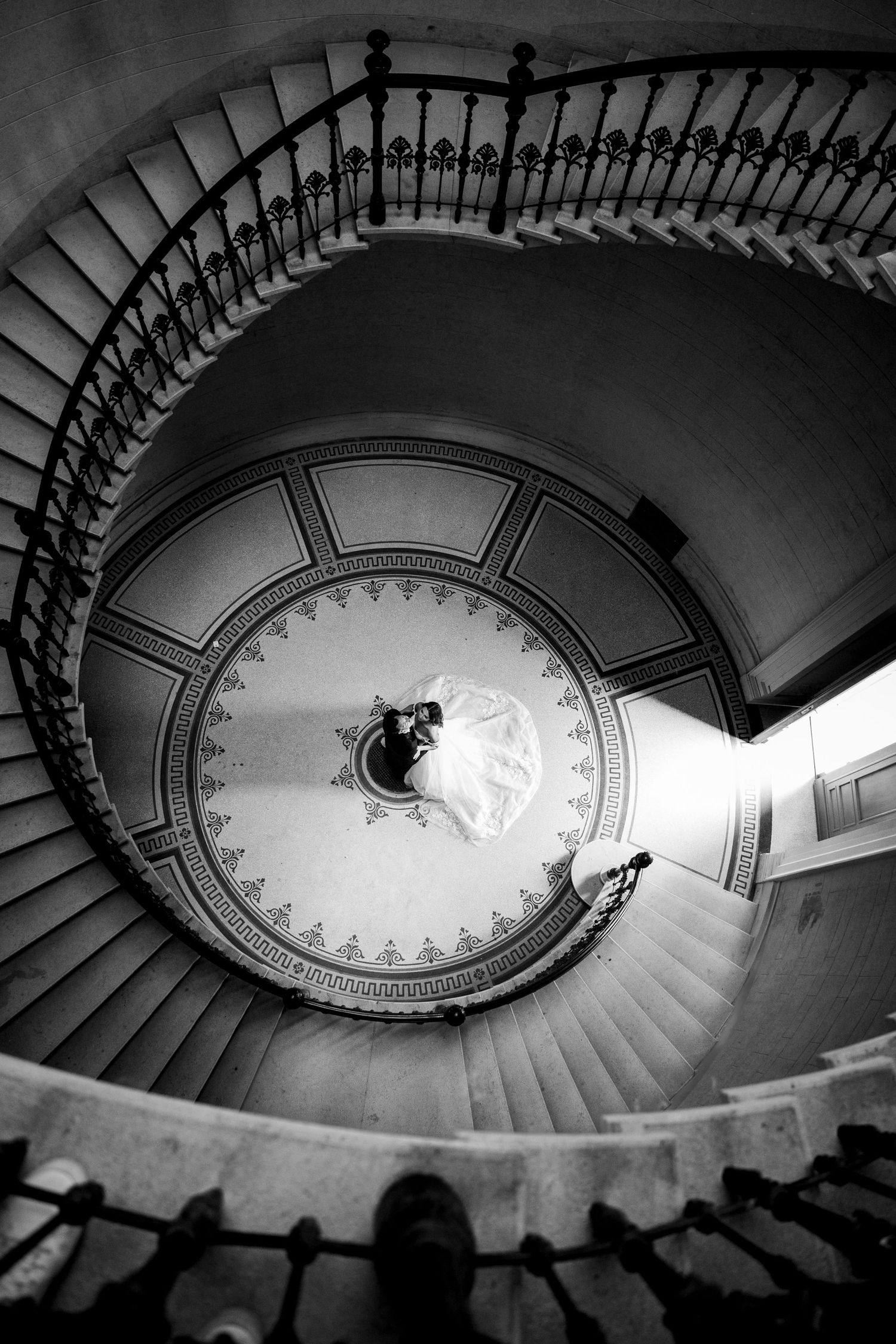 Aerial black and white view of bride and groom kissing at center of grand spiral staircase.