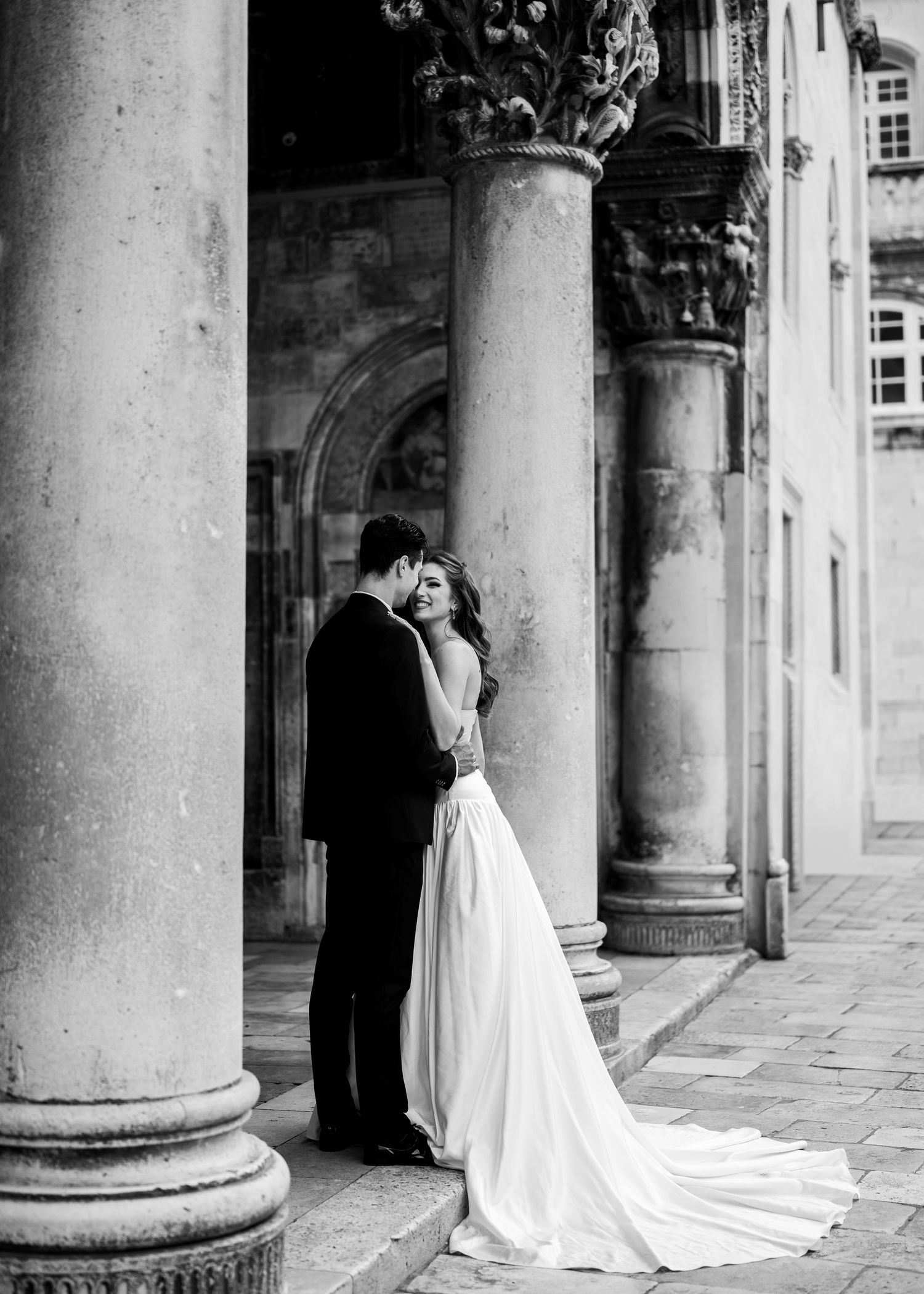Black and white romantic portrait of bride and groom embracing between classical stone columns of historic venue.