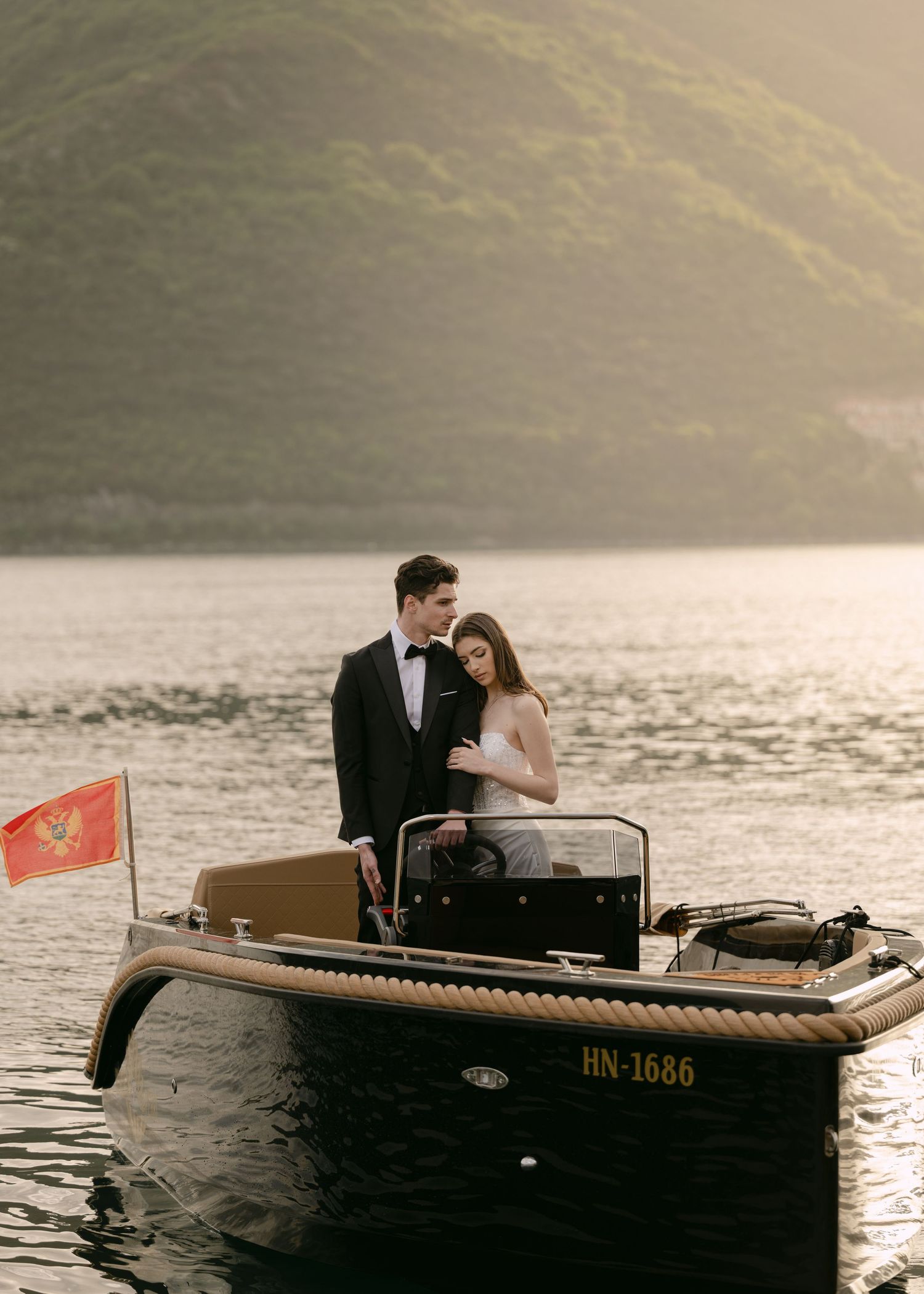 Elegant couple in wedding attire pose on a classic wooden boat on a scenic mountain lake.