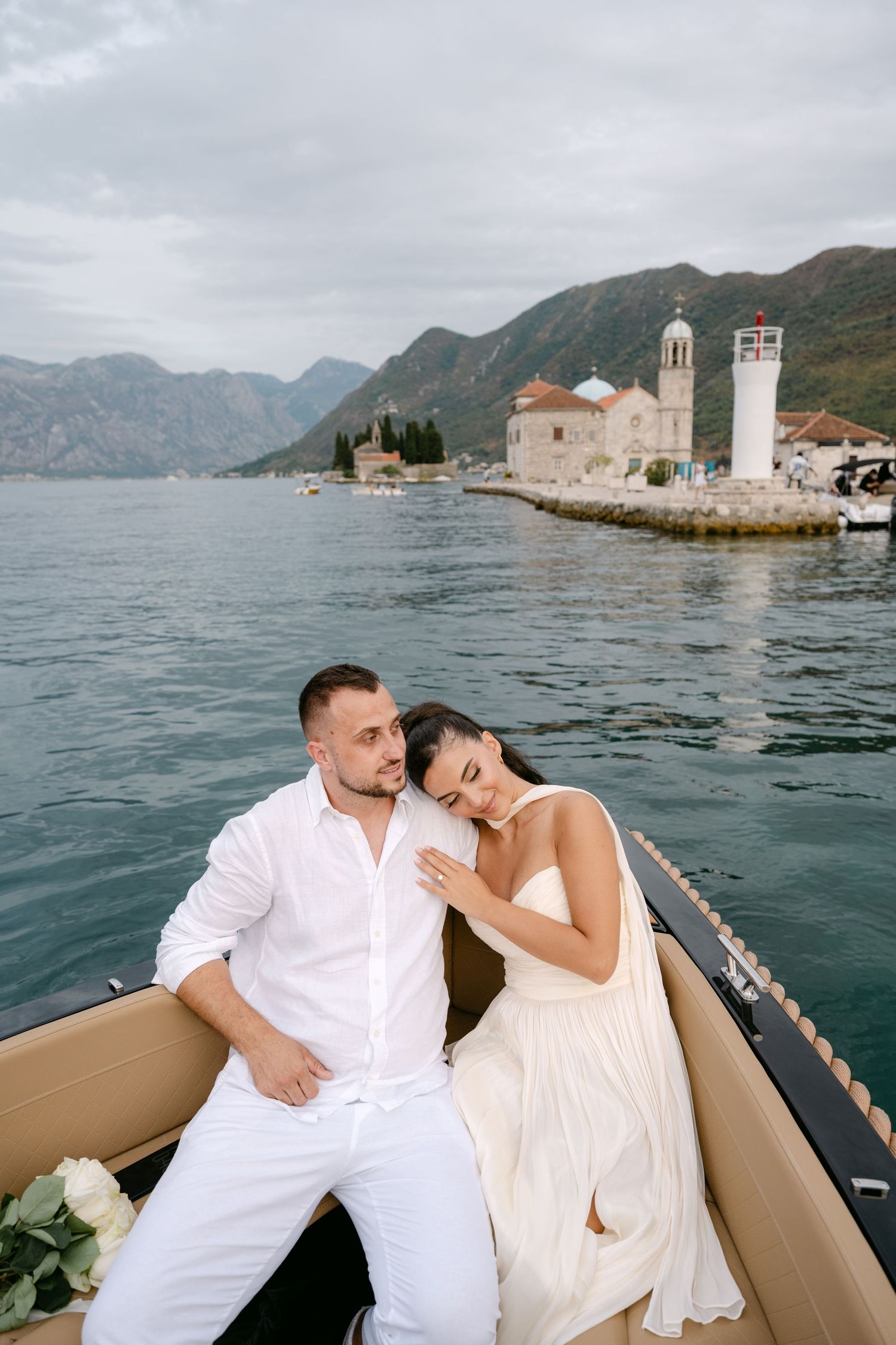 Romantic couple in white resting on a boat with Our Lady of the Rocks island church in Bay of Kotor.