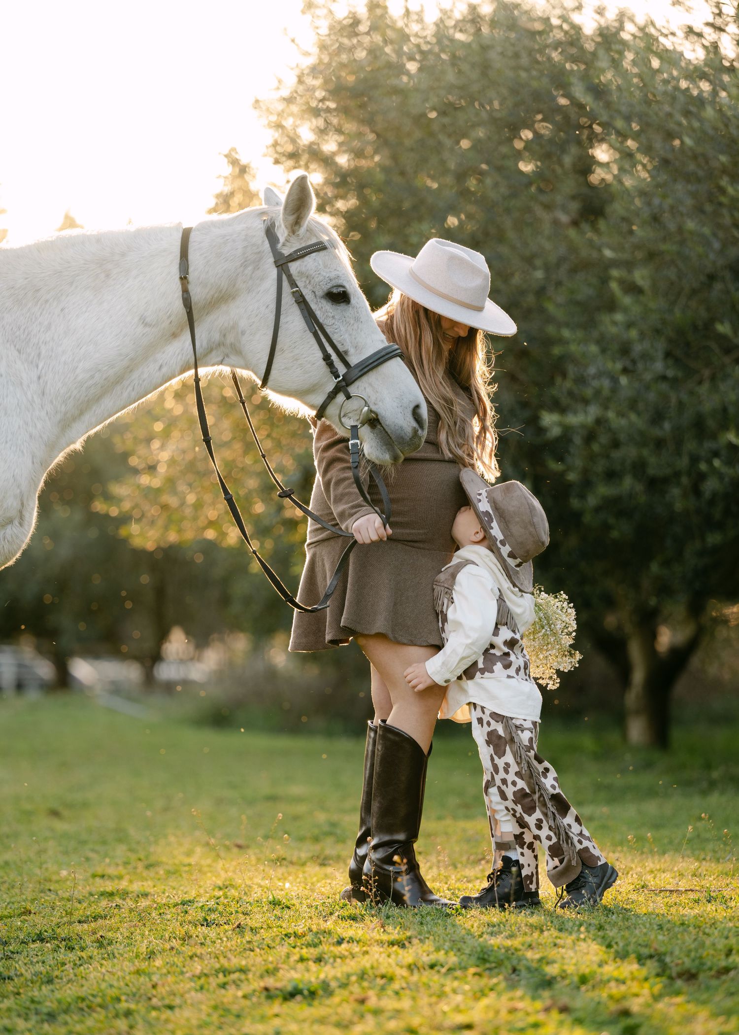Woman in wide-brim hat and tall boots with child in cowboy hat beside white horse at golden hour.
