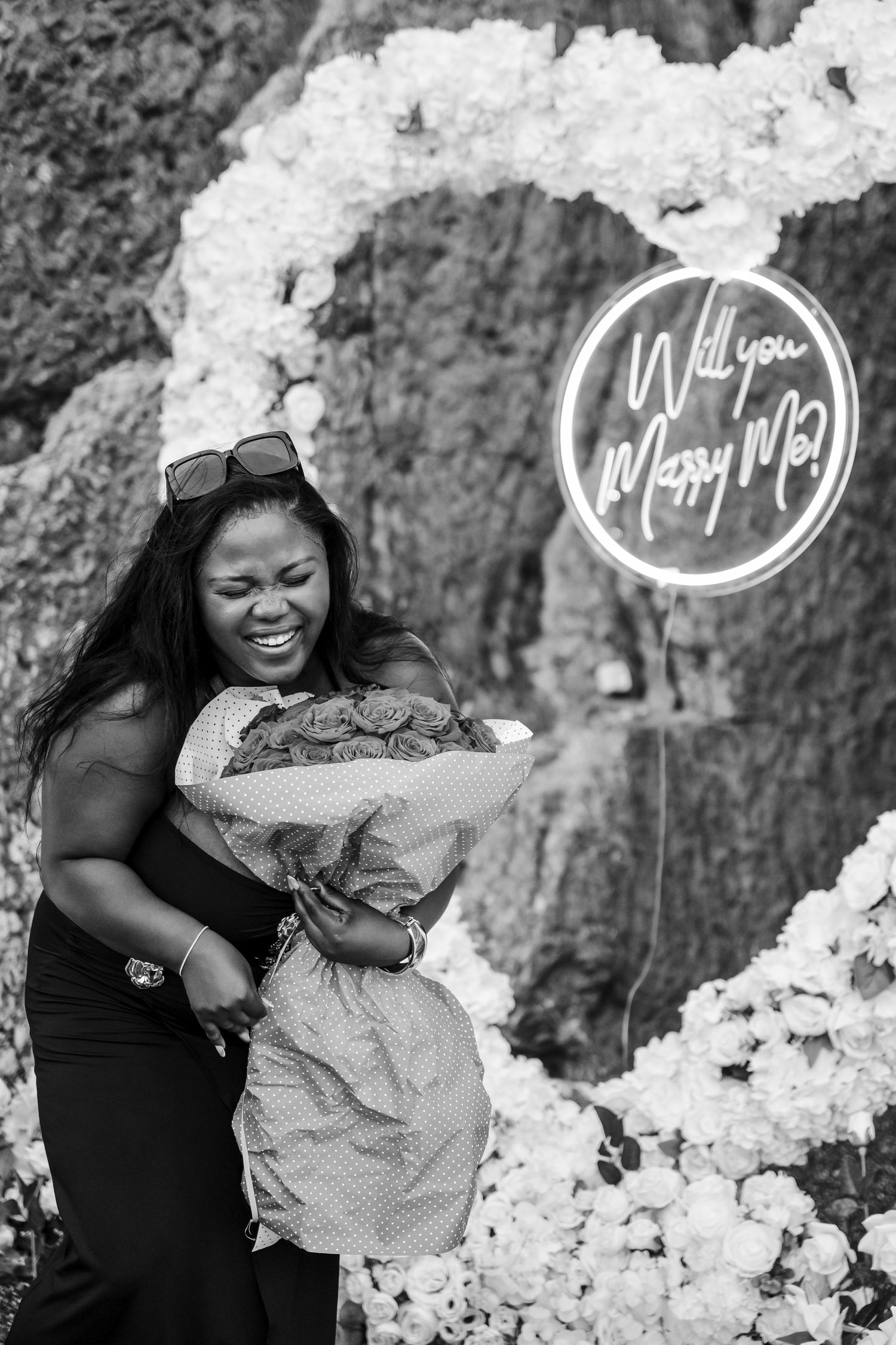 A joyful woman embraces her partner in front of a Will You Marry Me neon sign in black and white.