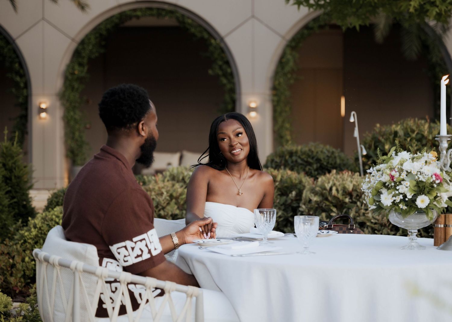 A couple holds hands and smiles at each other during a romantic candlelit outdoor dinner.