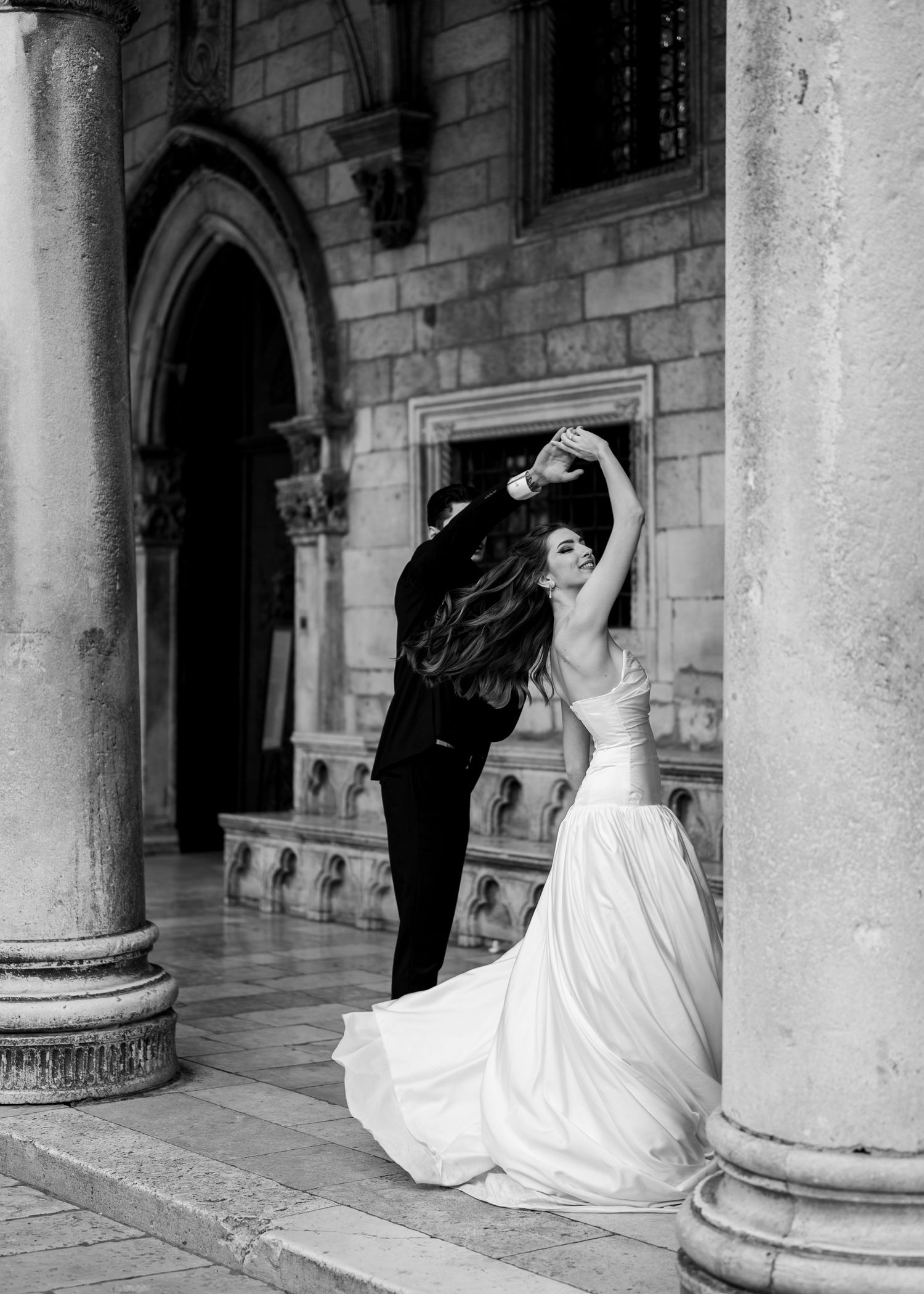 Bride and groom dancing elegantly between stone columns of a historic building in a black and white wedding photo.