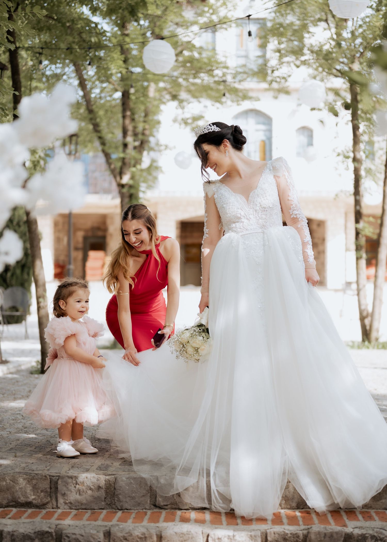 Bride in lace wedding gown with bridesmaid in red dress helping flower girl on stone steps outdoors.