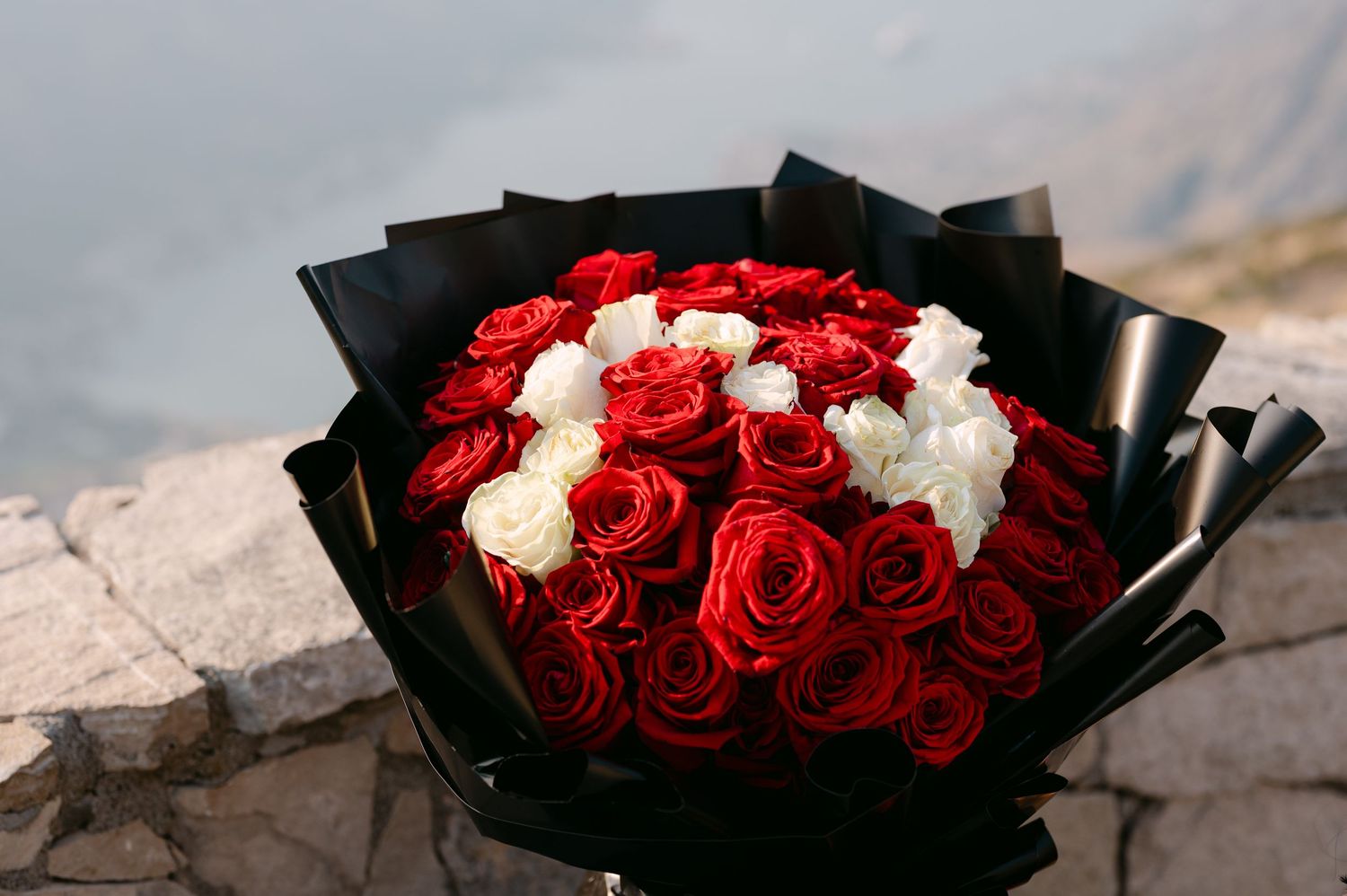 Elegant bouquet of red and white roses wrapped in black paper on a stone surface by the sea.