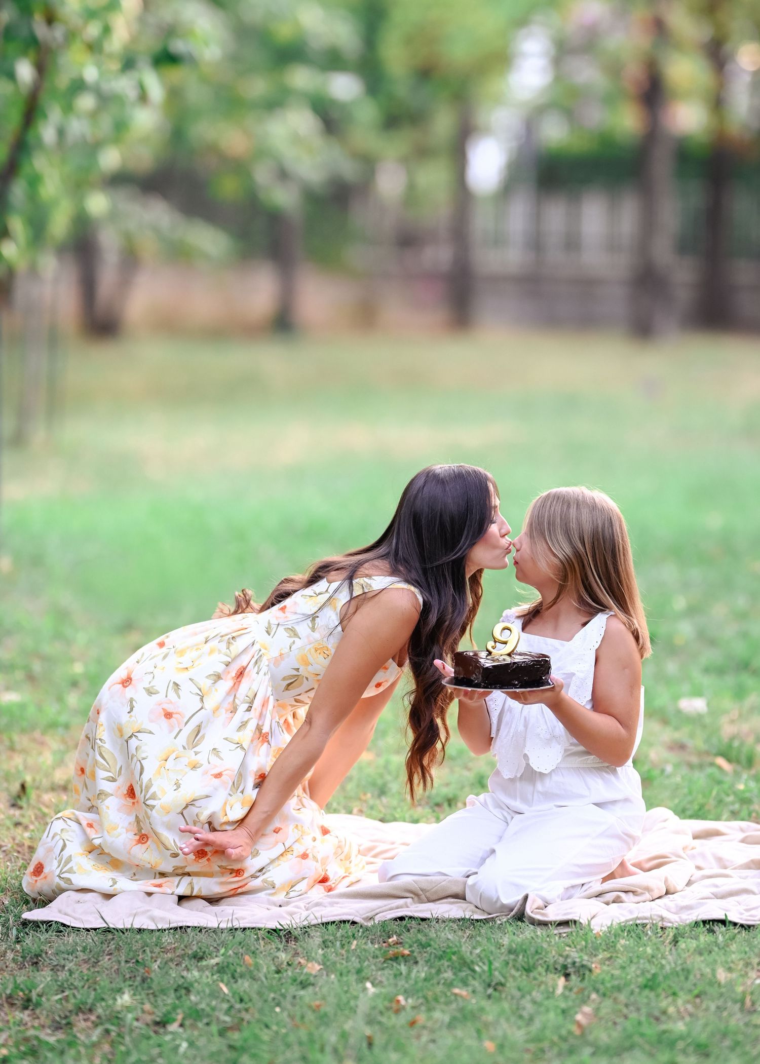Mother and daughter sharing a sweet kiss over birthday cake while sitting on grass together.