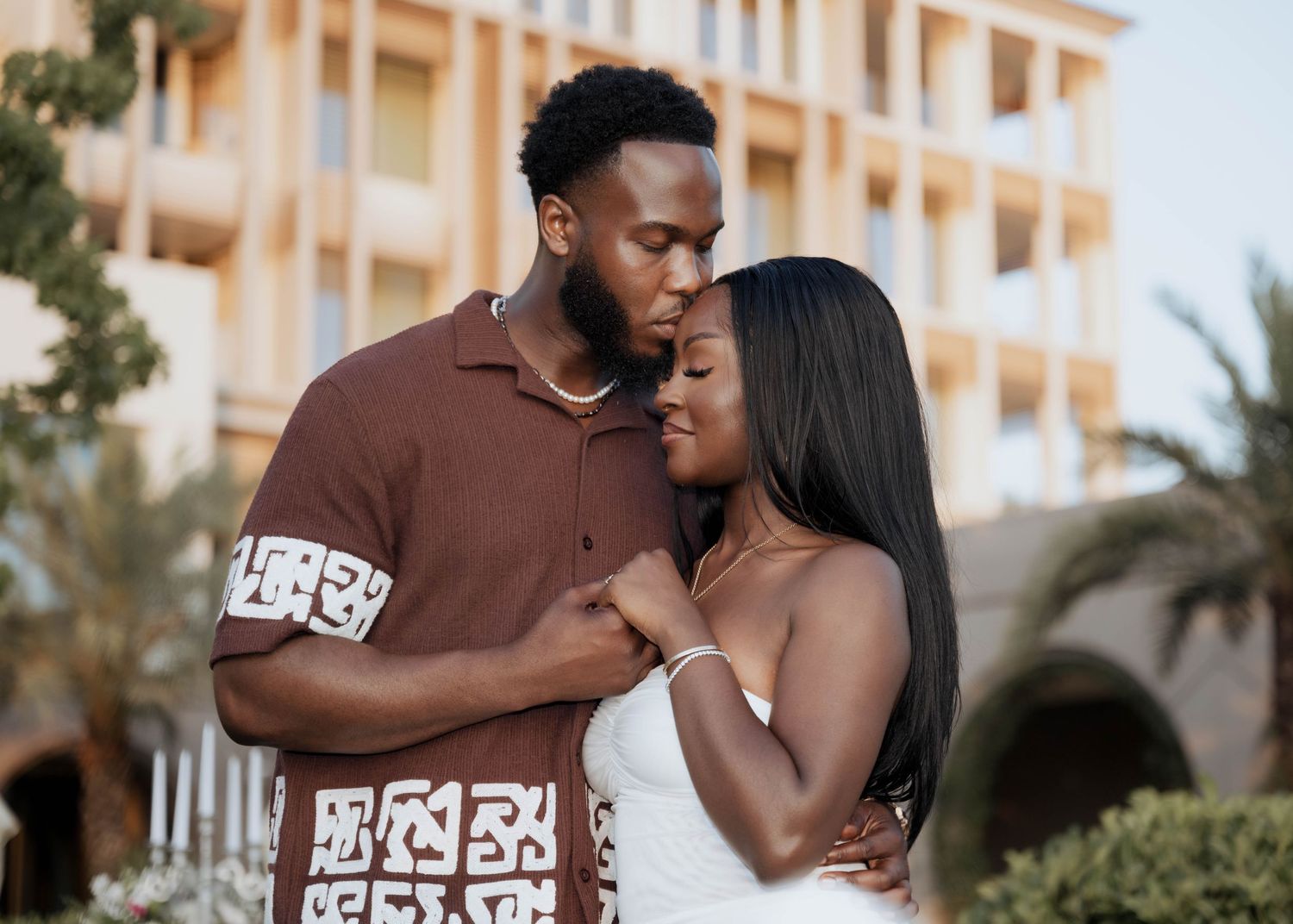 A couple shares a tender moment, the man kissing the womans forehead outdoors near a resort.