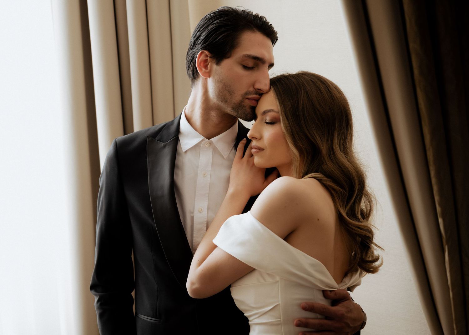 Groom in black suit tenderly embracing bride in off-shoulder white dress by curtained window.