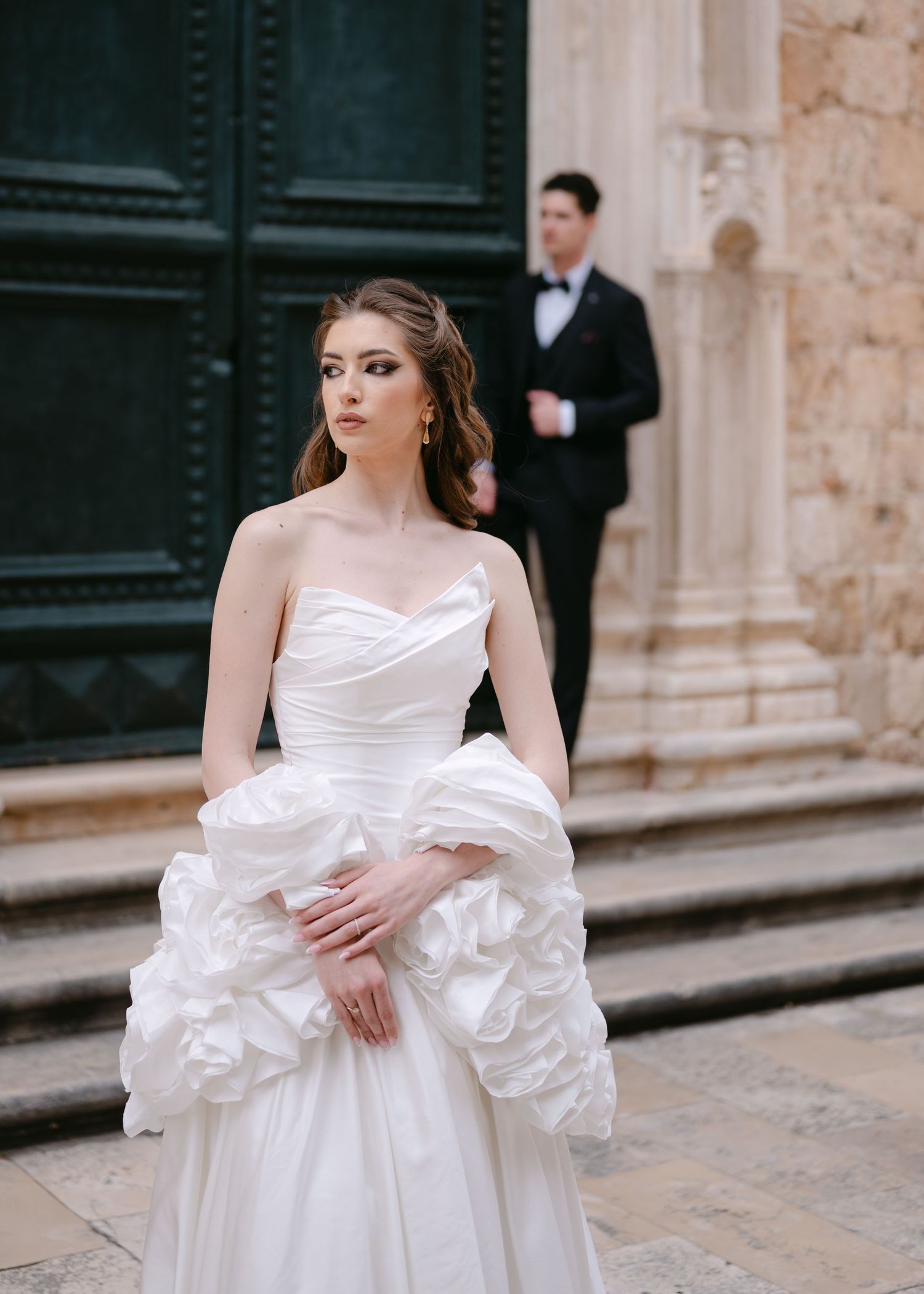 Elegant bride in strapless ruffled white gown gazes away while groom in tuxedo stands in background.