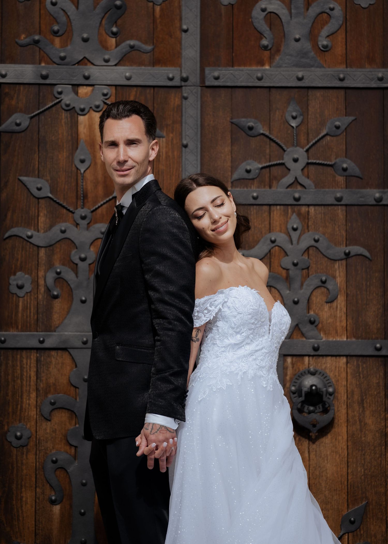 Bride and groom holding hands back-to-back before ornate carved wooden doors at historic venue.
