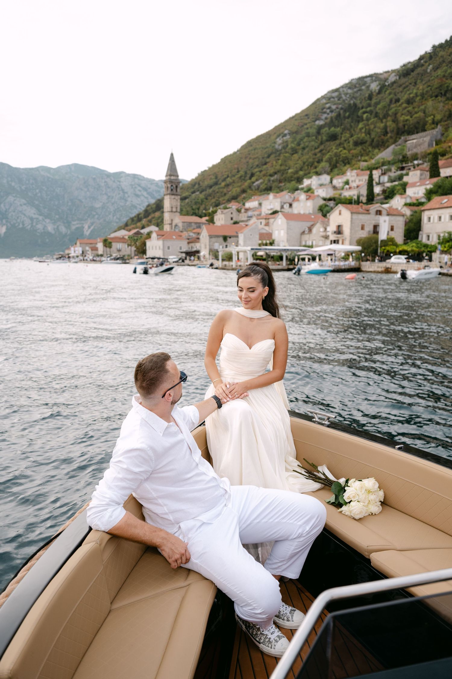 Bride in strapless gown and groom in white sitting on luxury boat with Perast village waterfront backdrop.