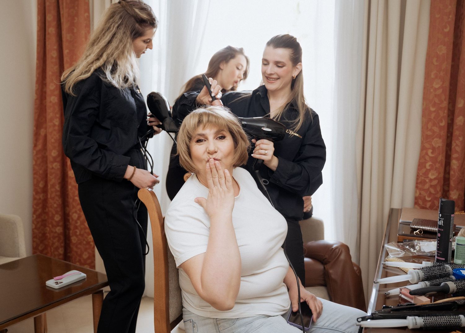 Woman getting hair styled by two hairdressers while blowing a kiss in bright room.