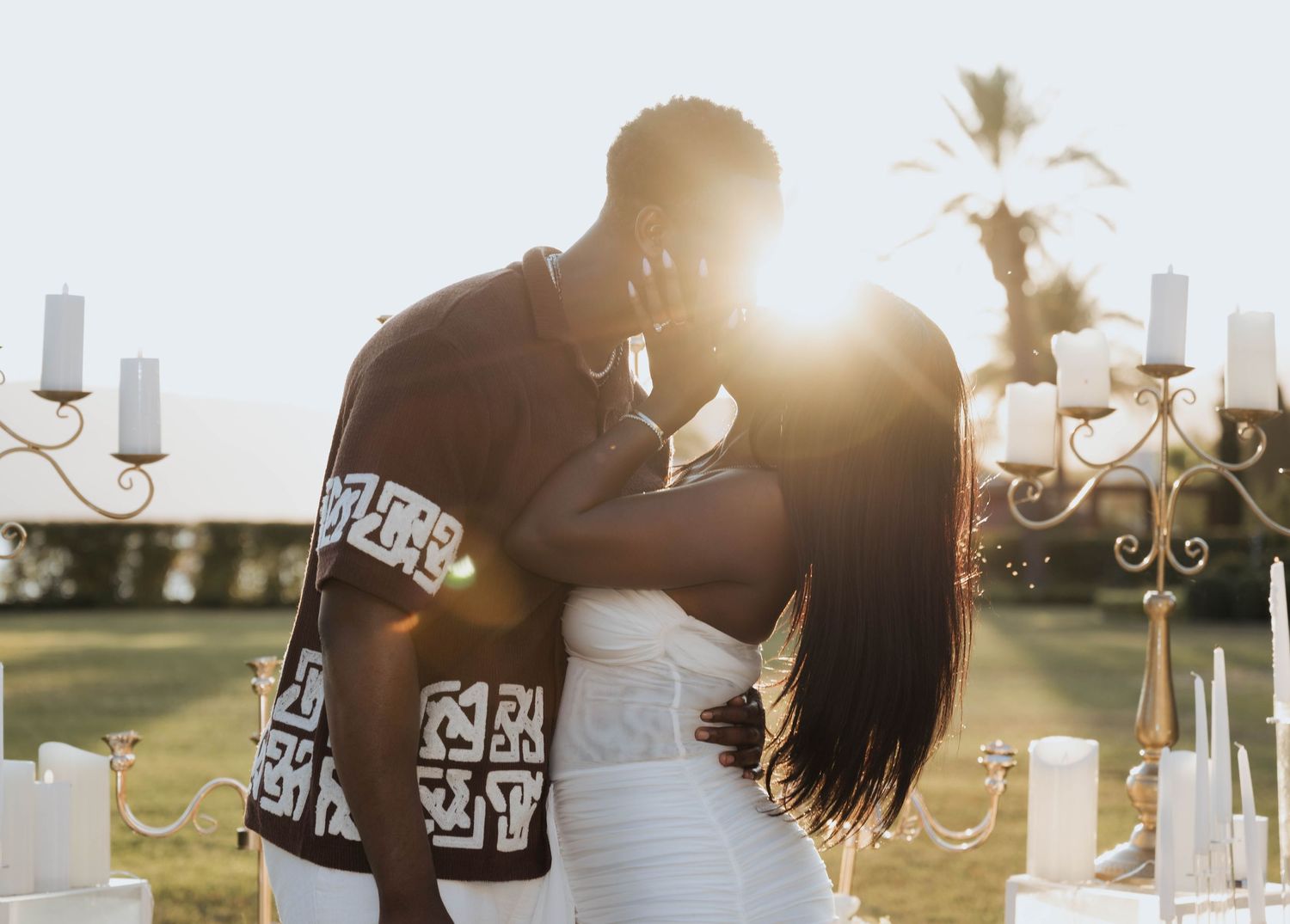 An engaged couple sharing a kiss backlit by golden sunset light at an outdoor venue.