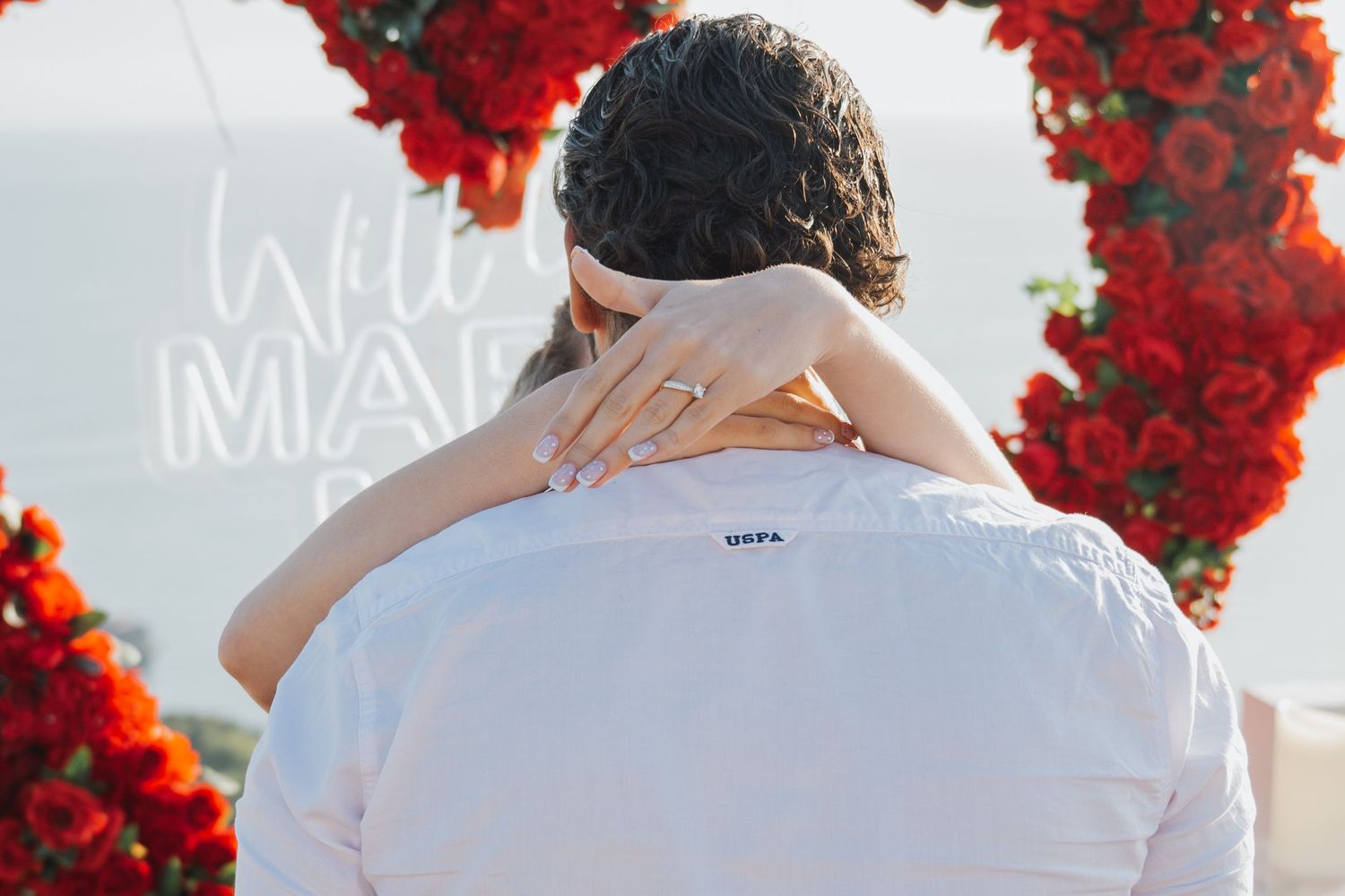 Couple embracing near red rose heart arch with 'Will You Marry Me' neon sign, engagement ring visible on hand.