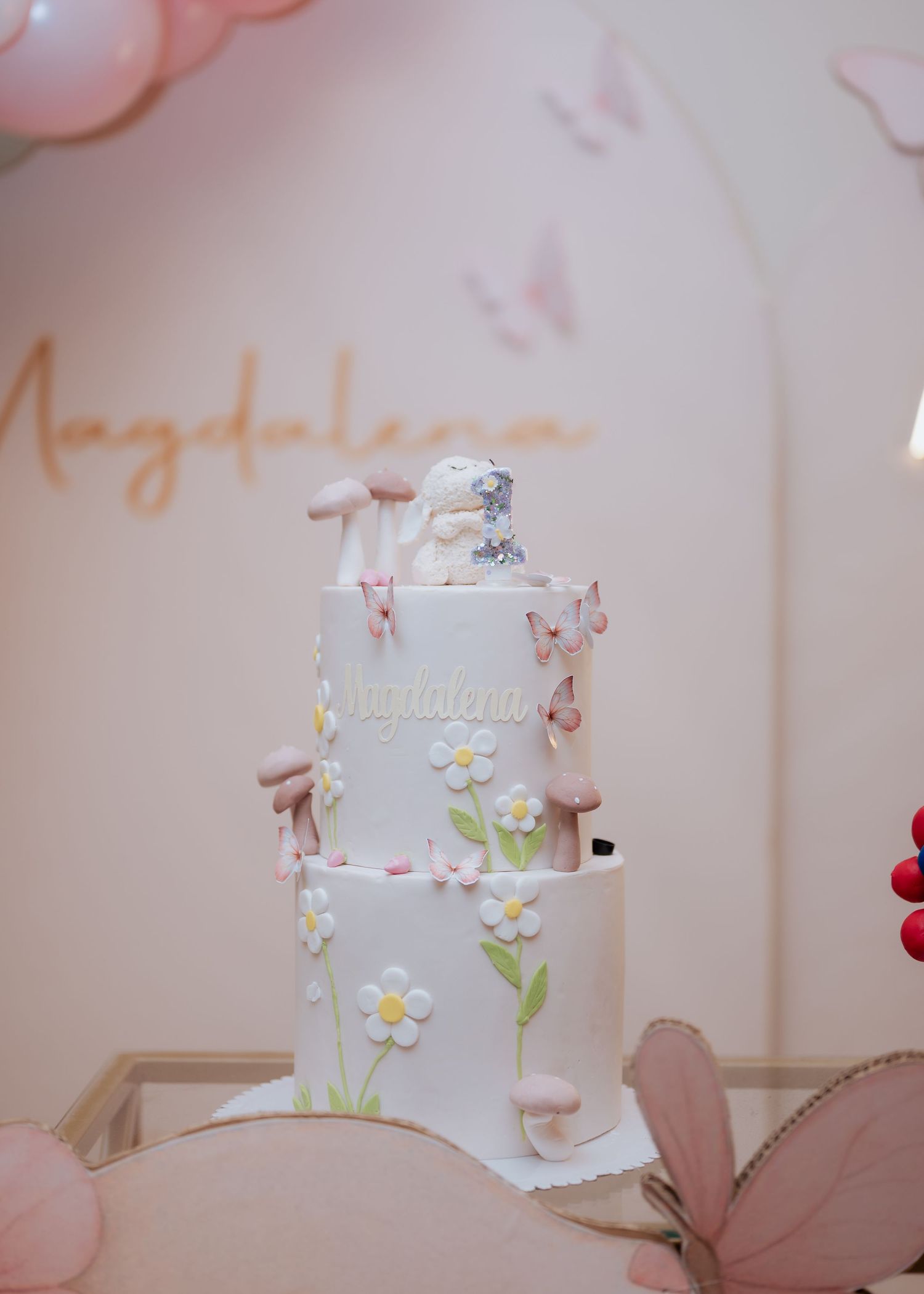 Elegant three-tier white birthday cake decorated with flowers, butterflies, and mushrooms reading Magdalena.