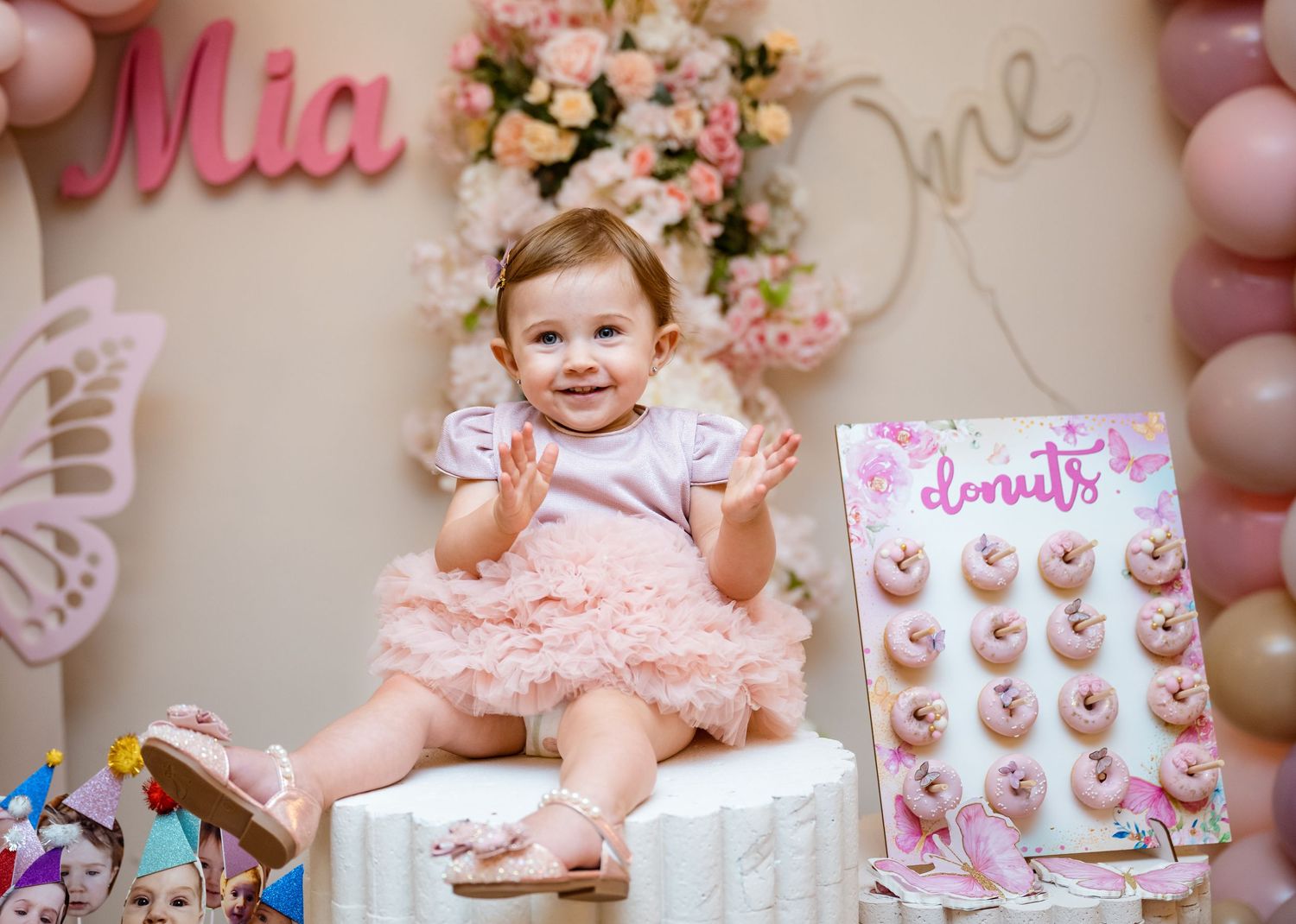 Smiling baby girl in pink tutu sitting beside a donut wall at her first birthday.