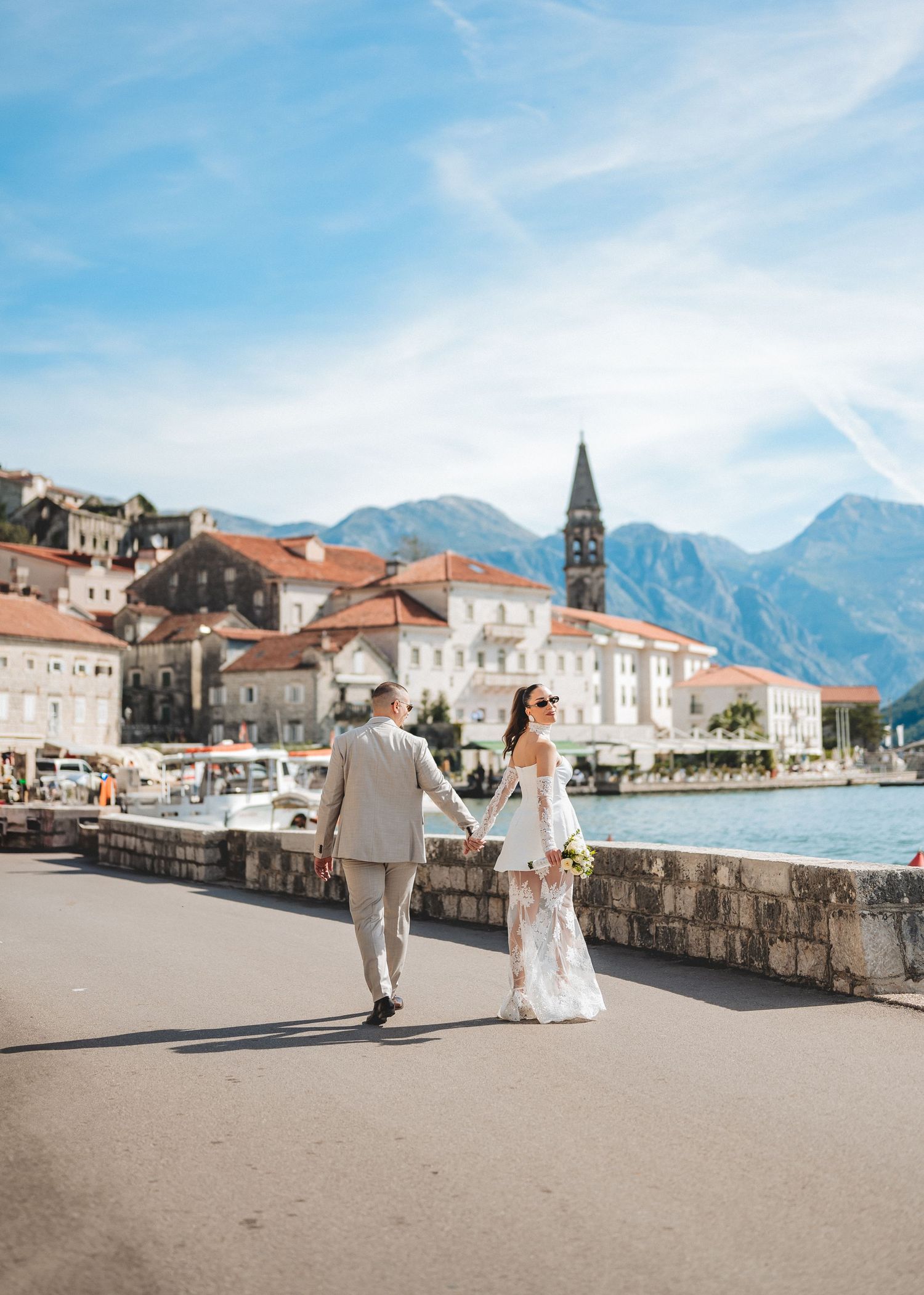 Bride and groom walking hand in hand along scenic Perast Montenegro waterfront on wedding day.