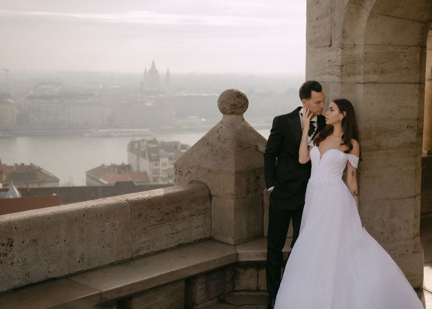 Wedding couple kissing on a stone balcony overlooking a scenic Budapest cityscape with river views.