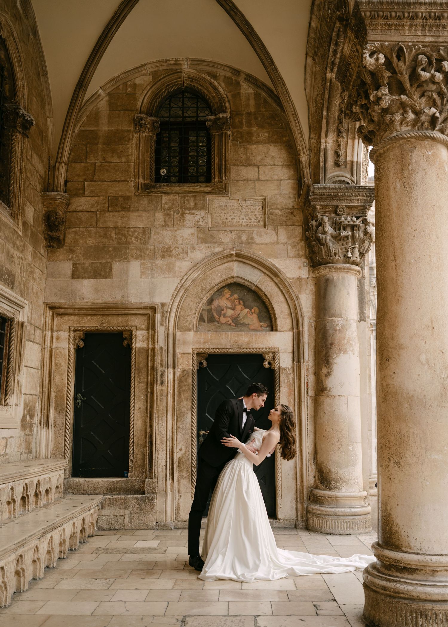 Bride and groom share romantic embrace in ornate historic stone archway with decorative columns and frescoes.
