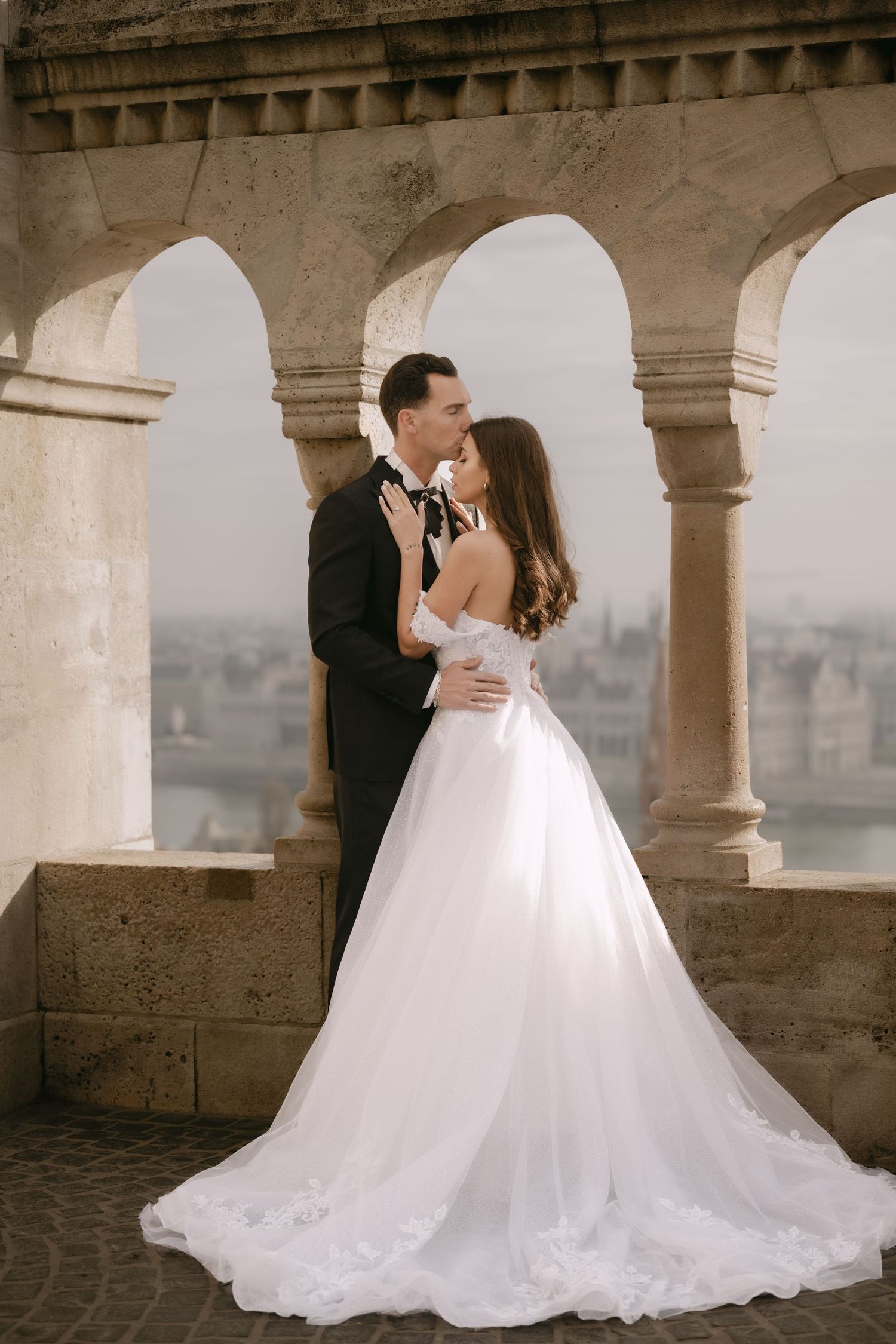 Bride in flowing white gown and groom embrace on stone balcony with arched columns and city view.