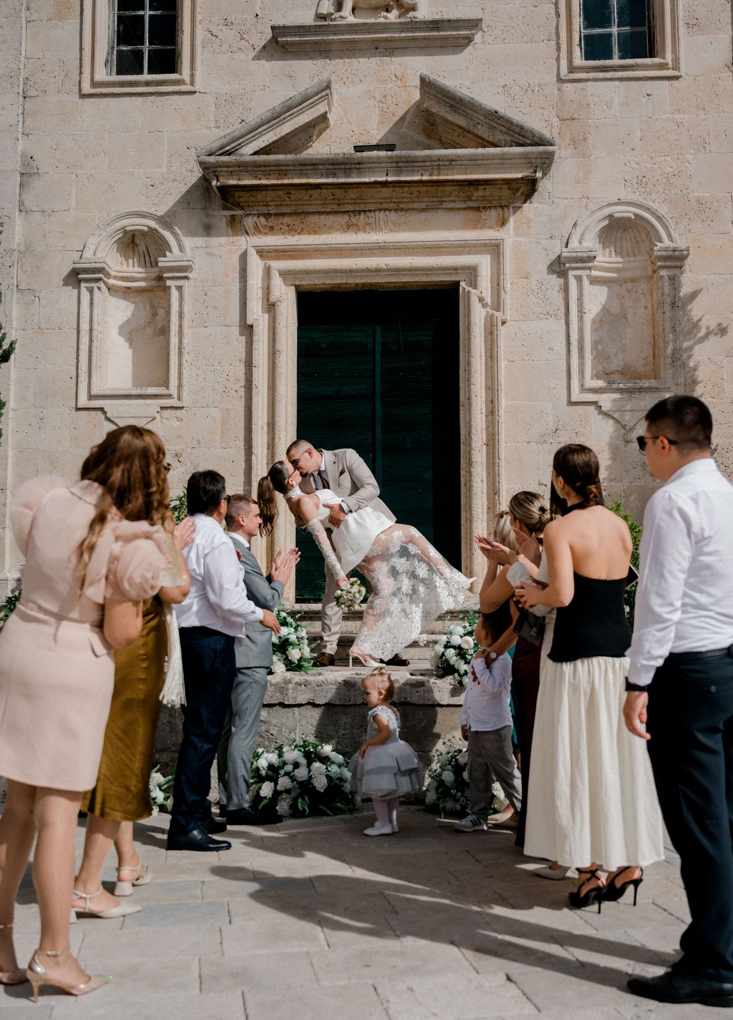 Bride and groom share a joyful kiss on church steps surrounded by celebrating wedding guests in a sunny European setting.