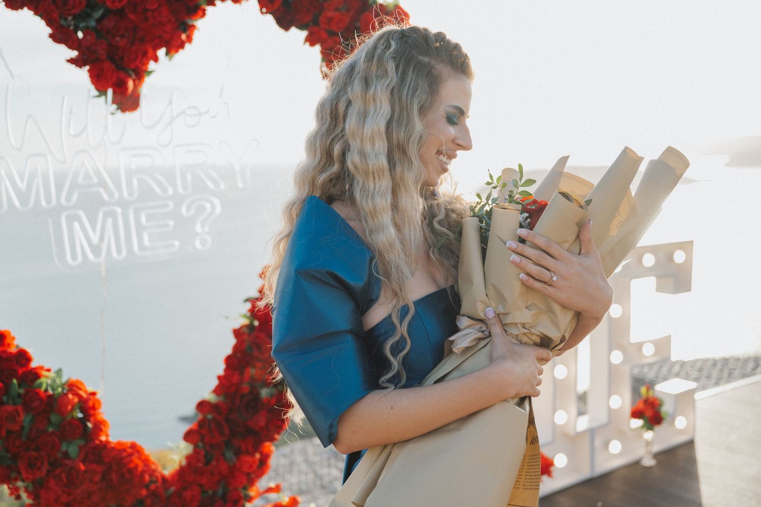 Smiling woman in blue dress holding bouquet near red rose heart arch with 'Will You Marry Me' neon sign.