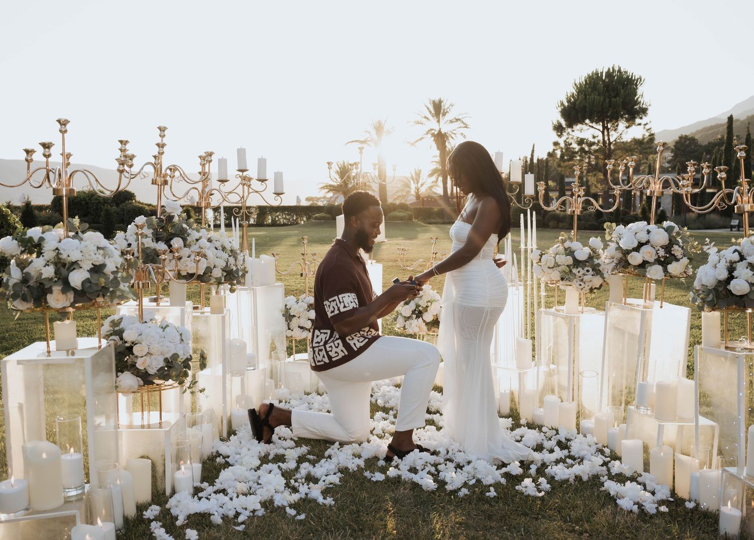 A romantic outdoor marriage proposal surrounded by white candles and floral arrangements at sunset.