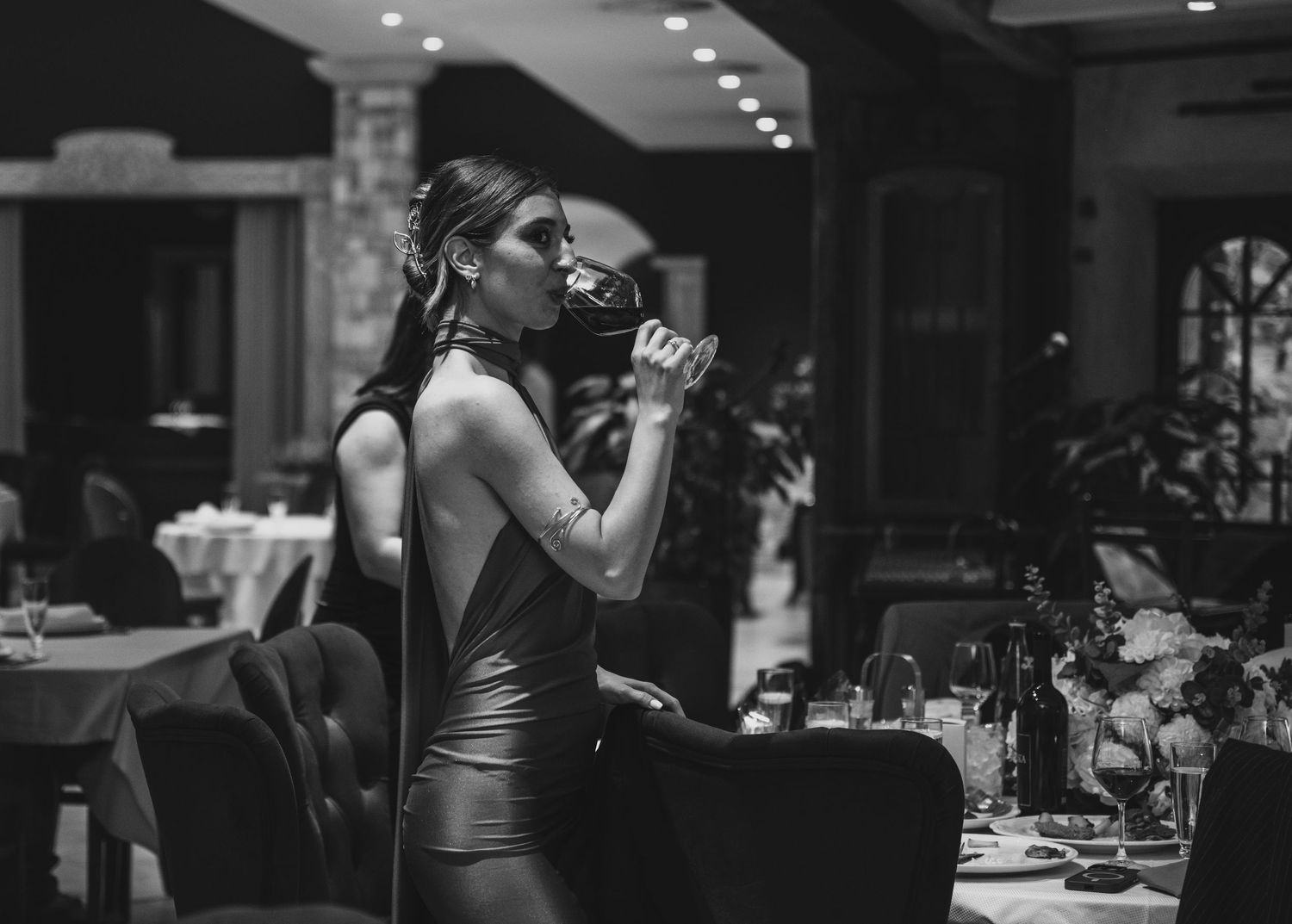 Elegant woman in backless dress sipping wine at upscale restaurant reception.