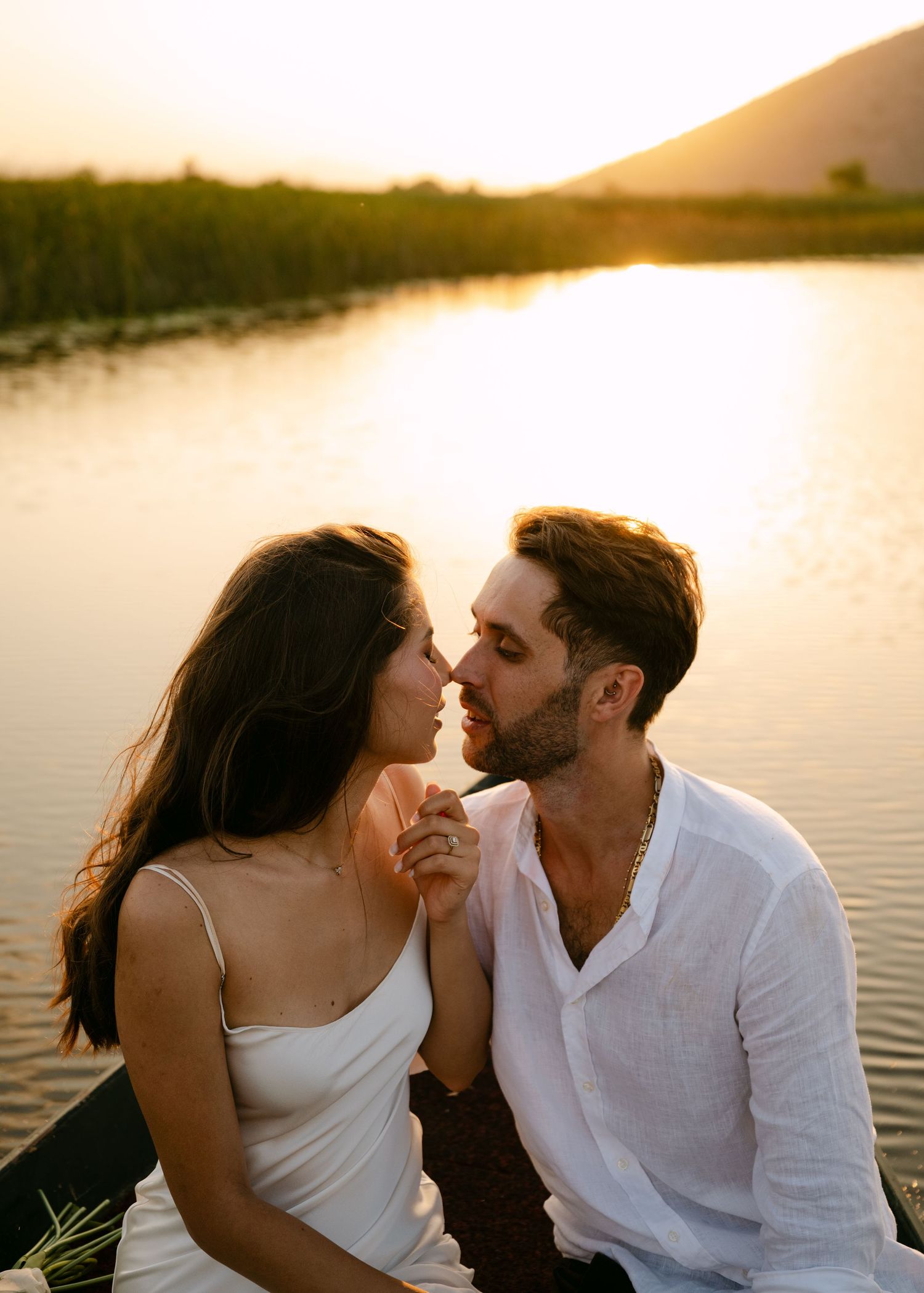 Couple sharing a romantic almost-kiss moment in a boat at golden sunset by a mountain lake.