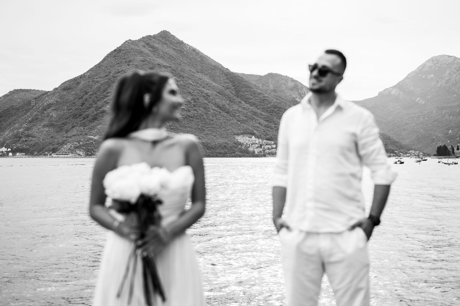 Black and white photo of bride and groom standing by the sea with mountain backdrop in Montenegro.