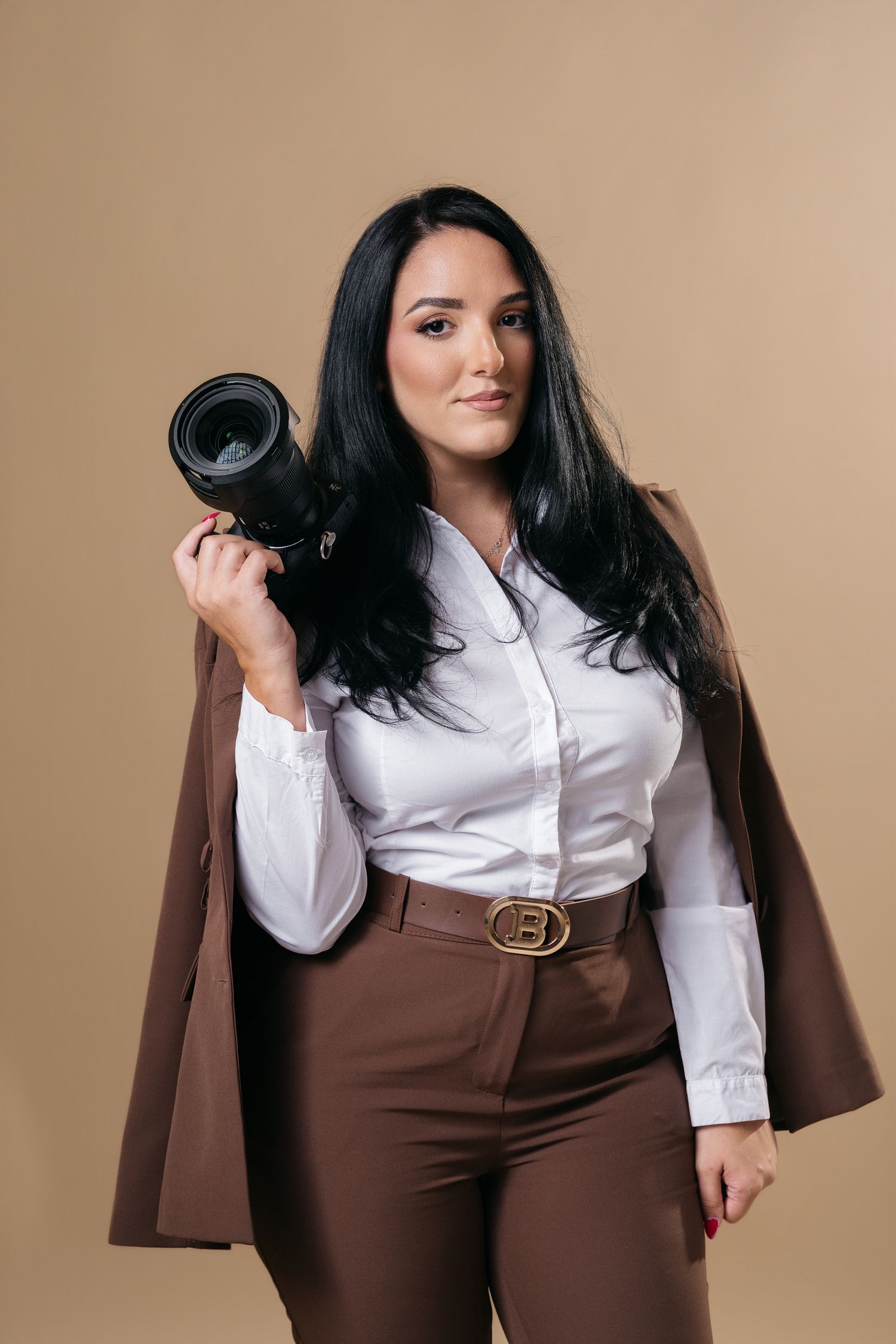 Professional female photographer in brown suit holds camera lens on beige studio background.