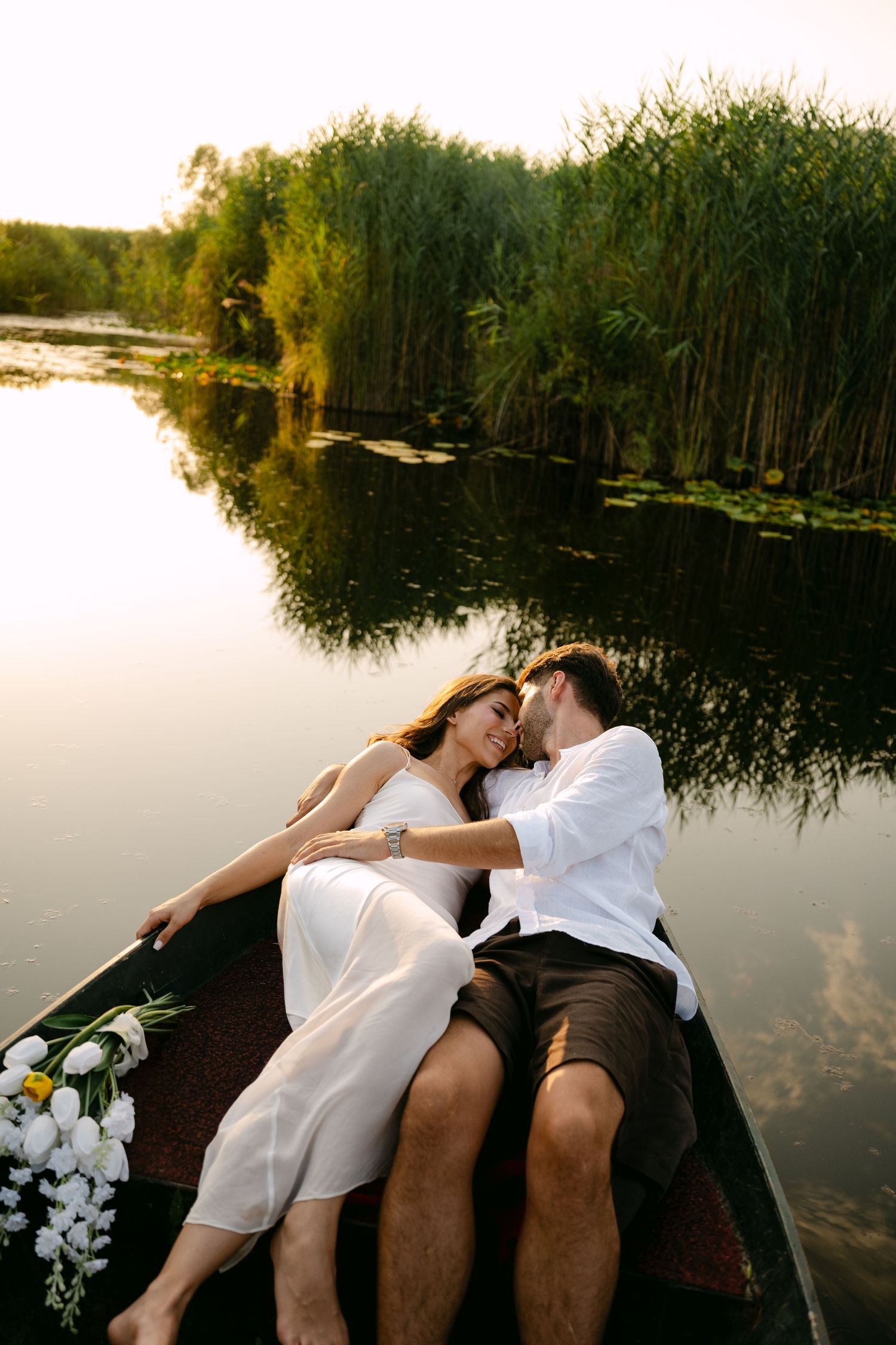 Romantic couple cuddling in a wooden boat surrounded by lush green reeds at sunset.